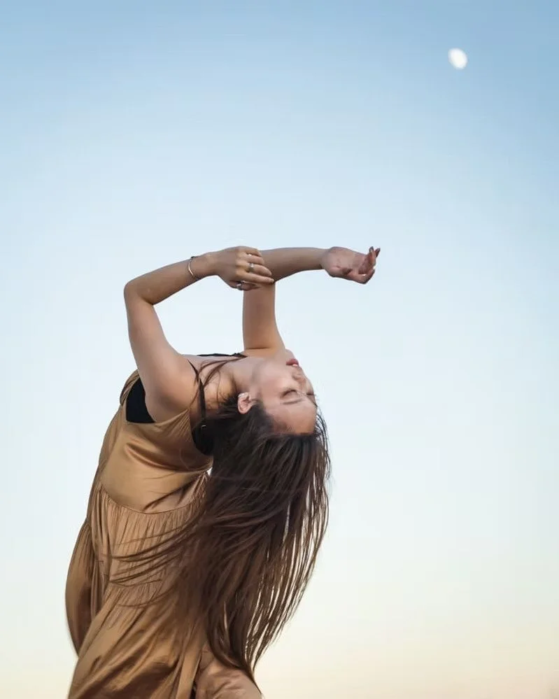 A woman with long hair and wearing a tan top is posing outdoors against a sky with the moon visible in the background.