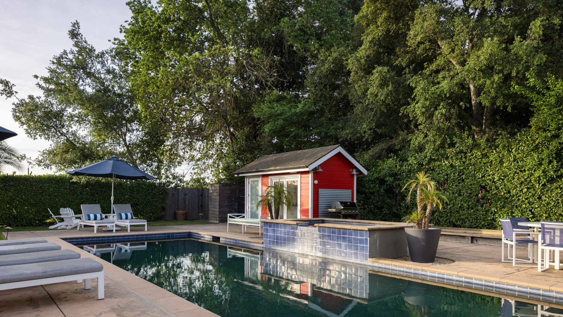 Backyard pool area with poolside lounge chairs, umbrellas, a small red pool house, potted plants, and lush green trees in the background.