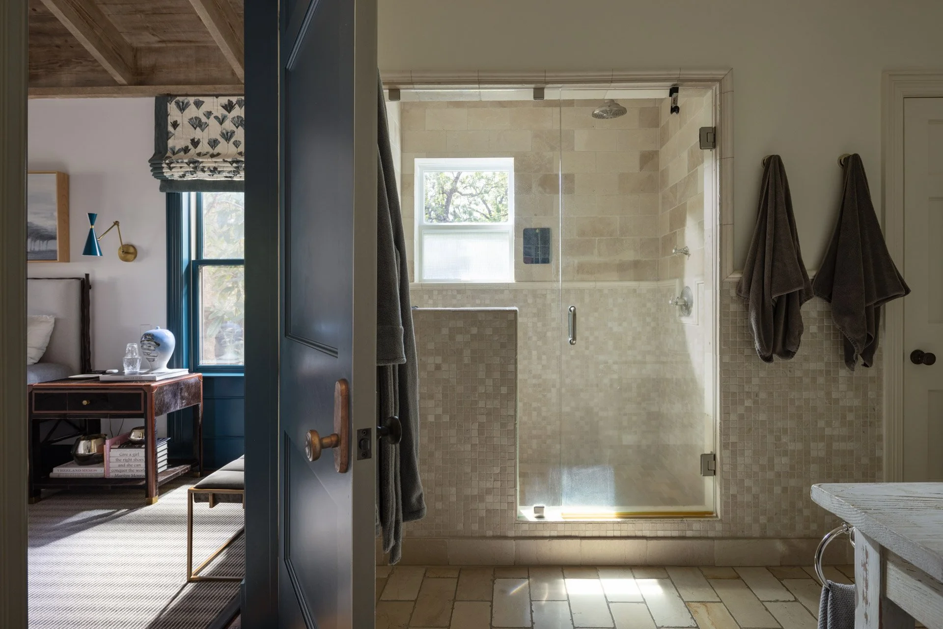 Door opening into a bedroom with a nightstand, lamp, and window, view into a bathroom with tiled shower and two hanging towels.