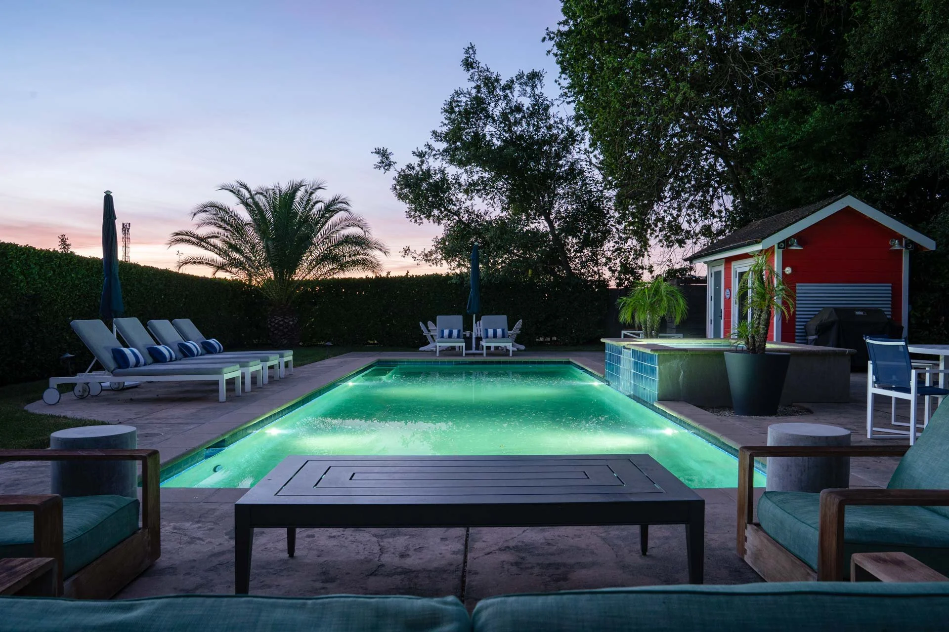 A backyard pool area illuminated at dusk with lounge chairs, umbrellas, a small red shed, and surrounding trees and shrubbery.