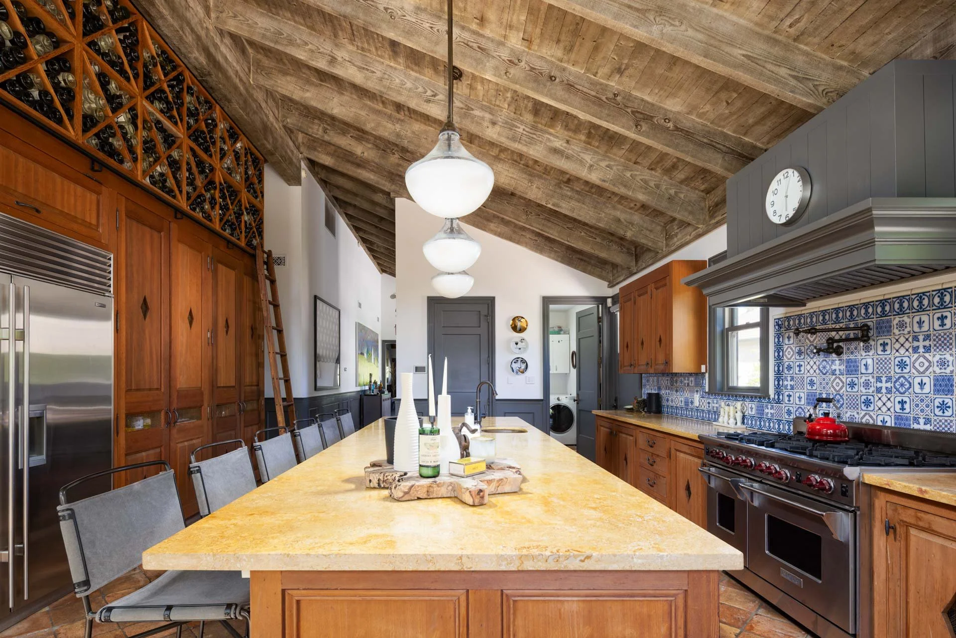A spacious kitchen with wooden cabinets, a large yellow marble island, stainless steel appliances, and a decorative blue and white tile backsplash. Wooden ceiling with pendant lighting and a wine storage area overhead.