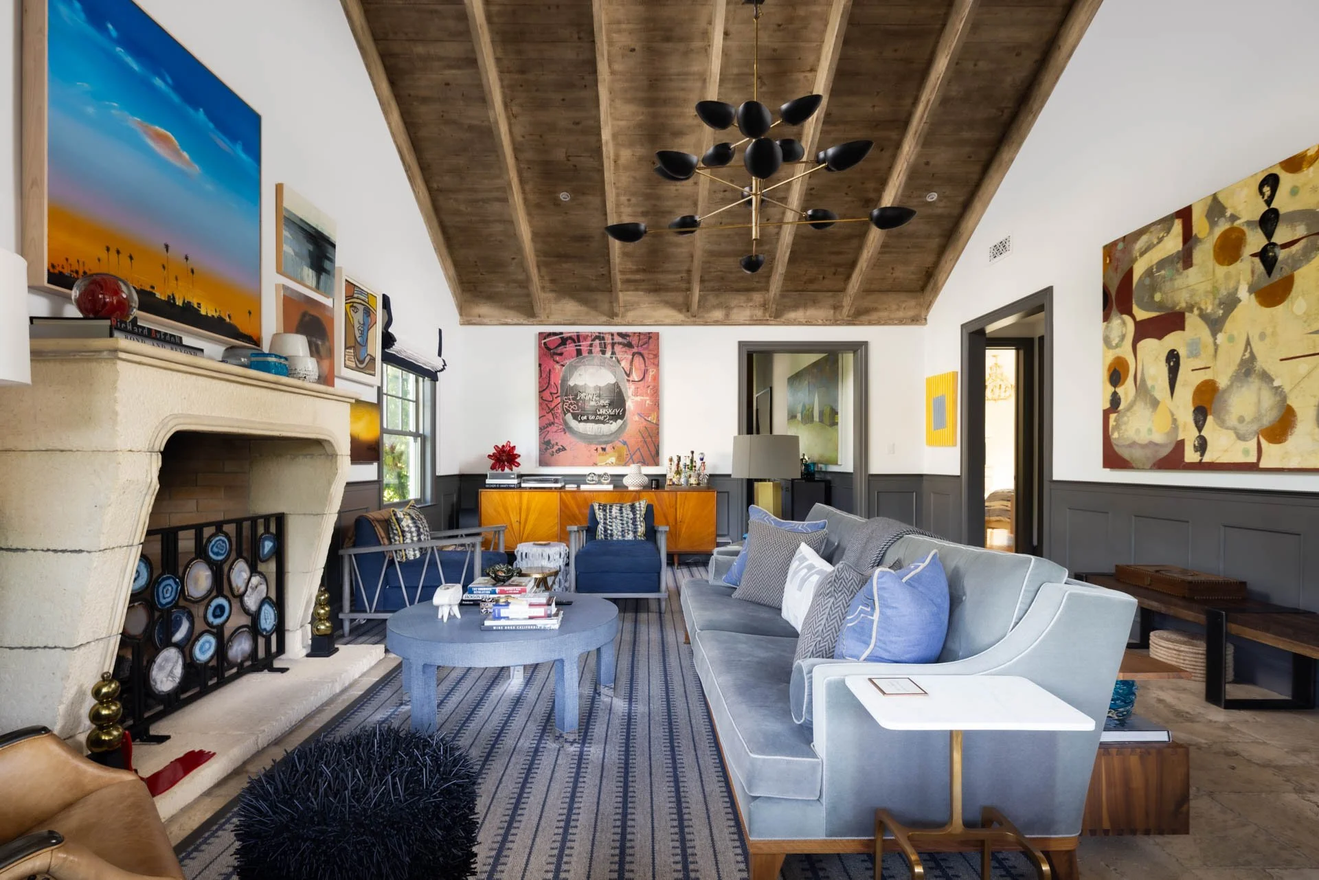 Spacious living room with a wooden ceiling, white walls with gray wainscoting, and a mix of colorful artwork. Features a stone fireplace, light blue sofa with pillows, a round blue coffee table, and various chairs. Decor includes books, lamps, and ar