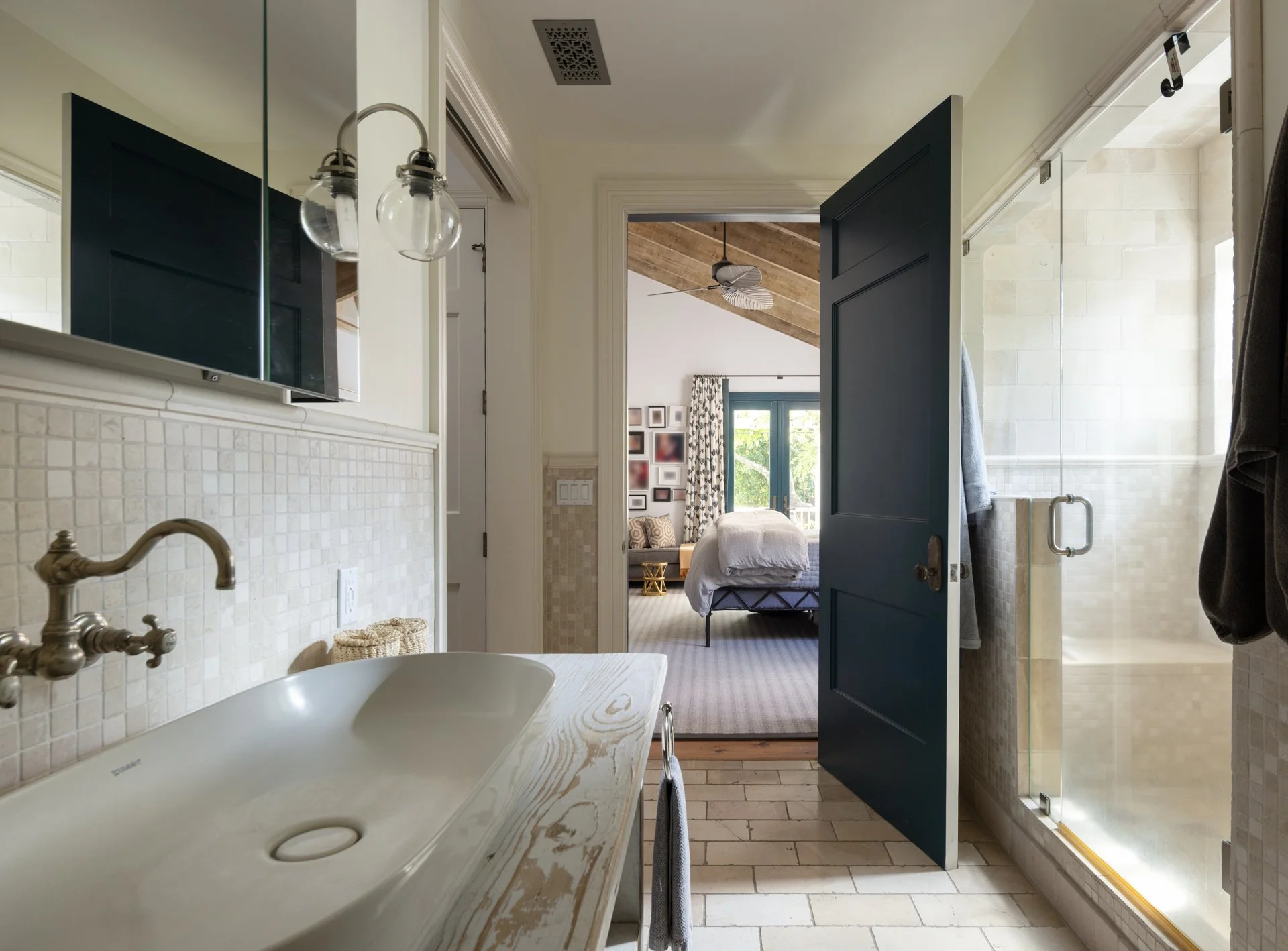 View from a bathroom looking into a bedroom through an open door. The bathroom has a beige sink, beige tiled walls, and a glass shower door. The bedroom has a bed with white bedding, a ceiling fan, a window with curtains, and wall art. The ceiling in