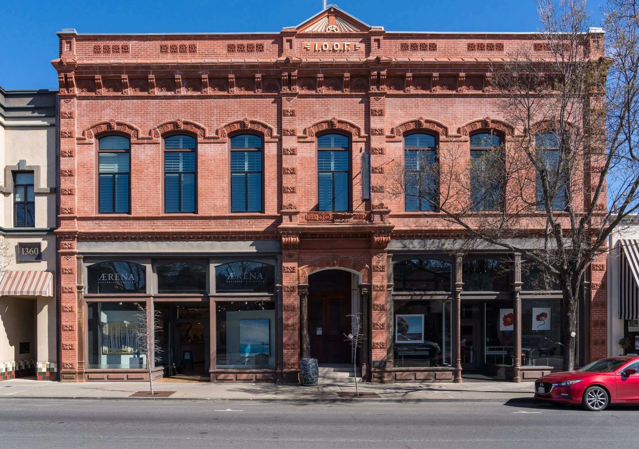 A three-story red brick historic building with arched windows on the upper floors. The ground floor has large glass display windows for art galleries labeled "AERENA GIF HALL". There are two leafless trees in front, with a red car parked on the stree