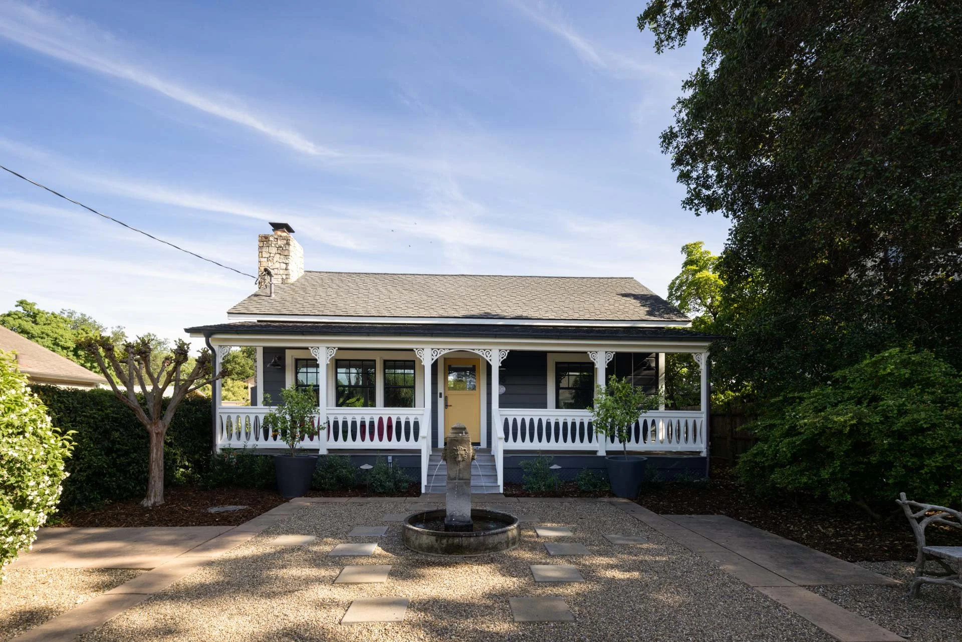 A house with a gray exterior, front porch with white decorative trim, yellow front door, and a small fountain in the front yard. There are trees and bushes around, with a clear blue sky overhead.