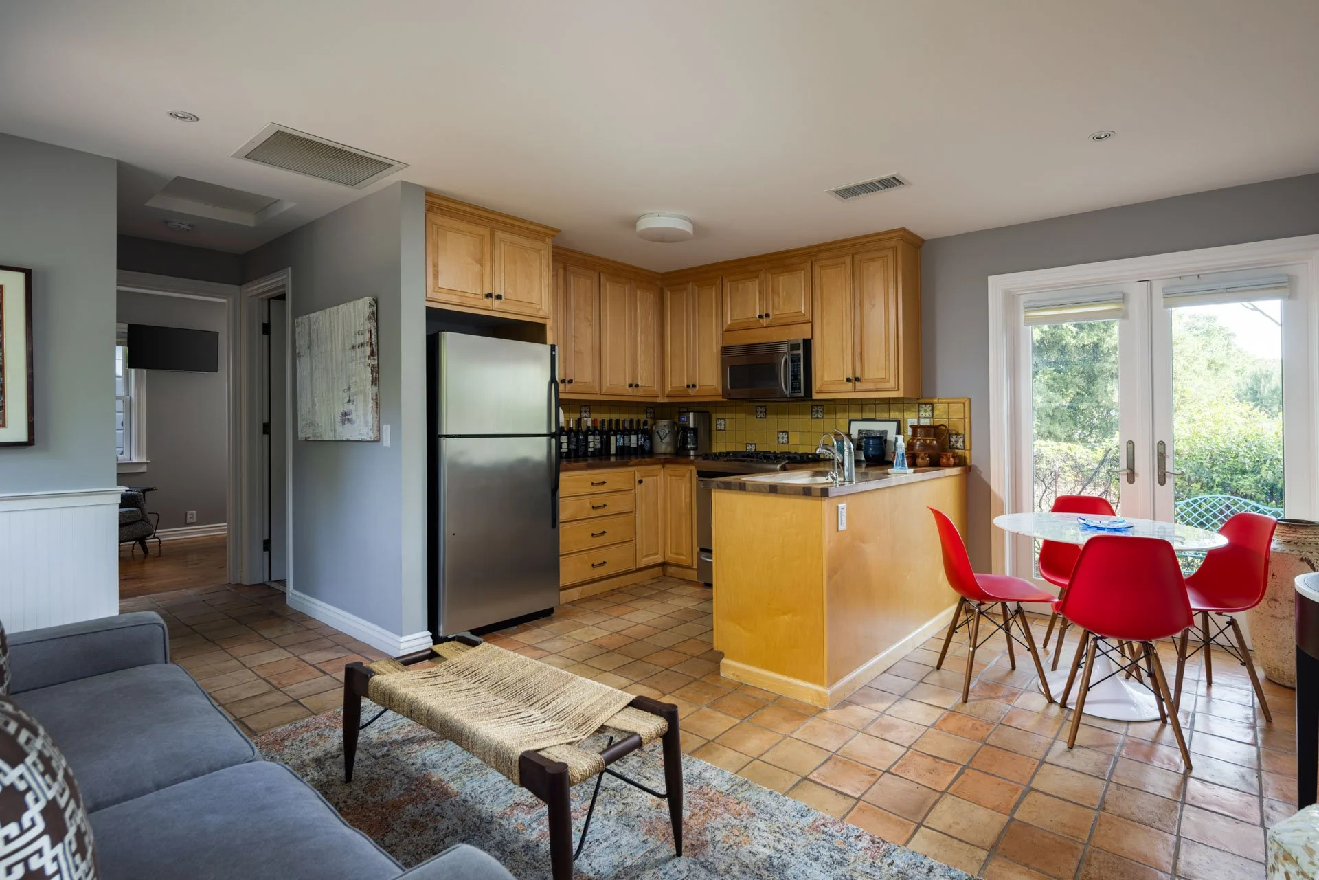 Open concept kitchen and dining area with gray walls, wooden cabinets, yellow tile backsplash, stainless steel refrigerator, and a round glass table with four red chairs, leading to outdoor scenery through glass doors.