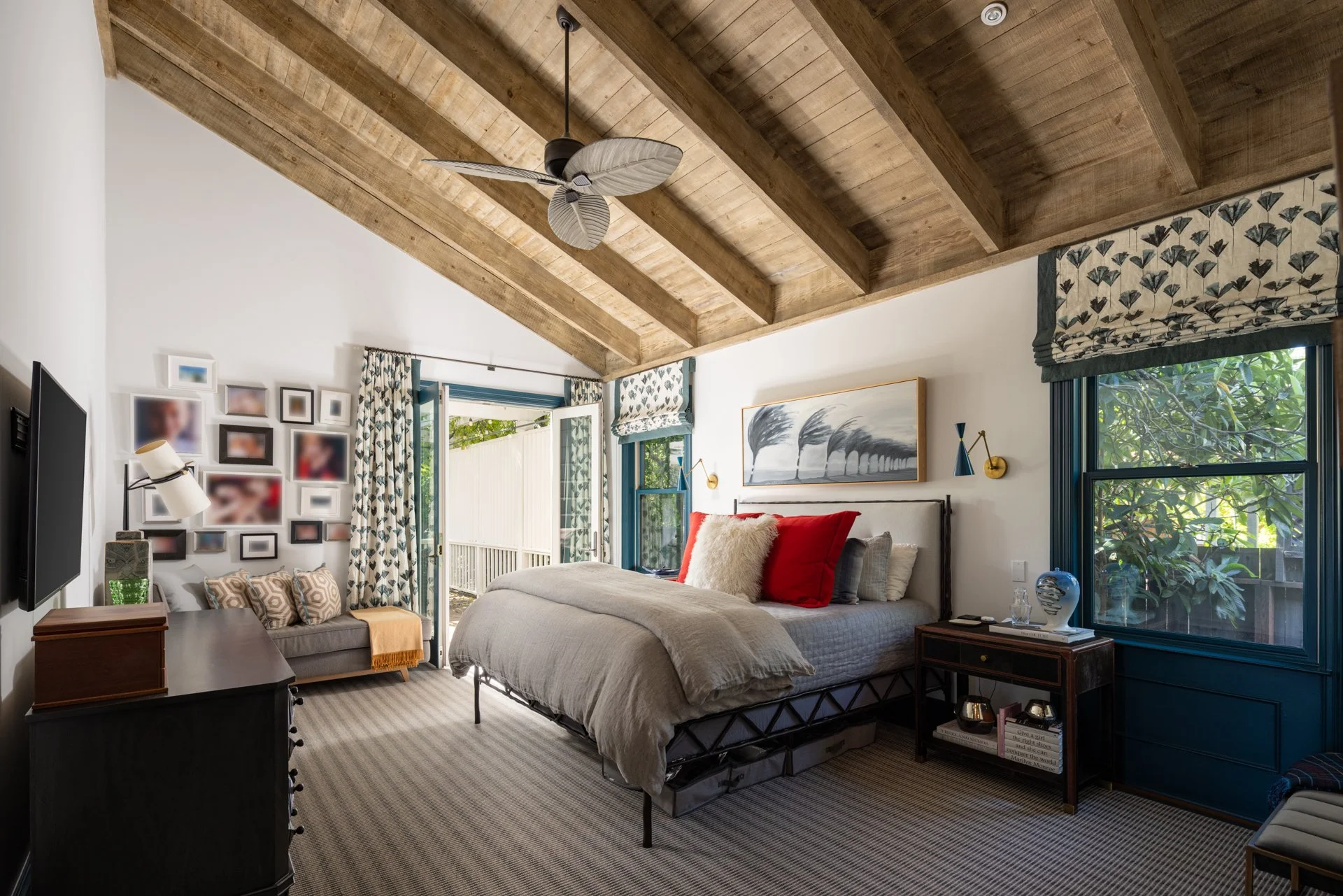 A cozy bedroom with a wooden vaulted ceiling, a large bed with multiple pillows, a small sofa, a side table with a decorative vase, and windows with patterned curtains.