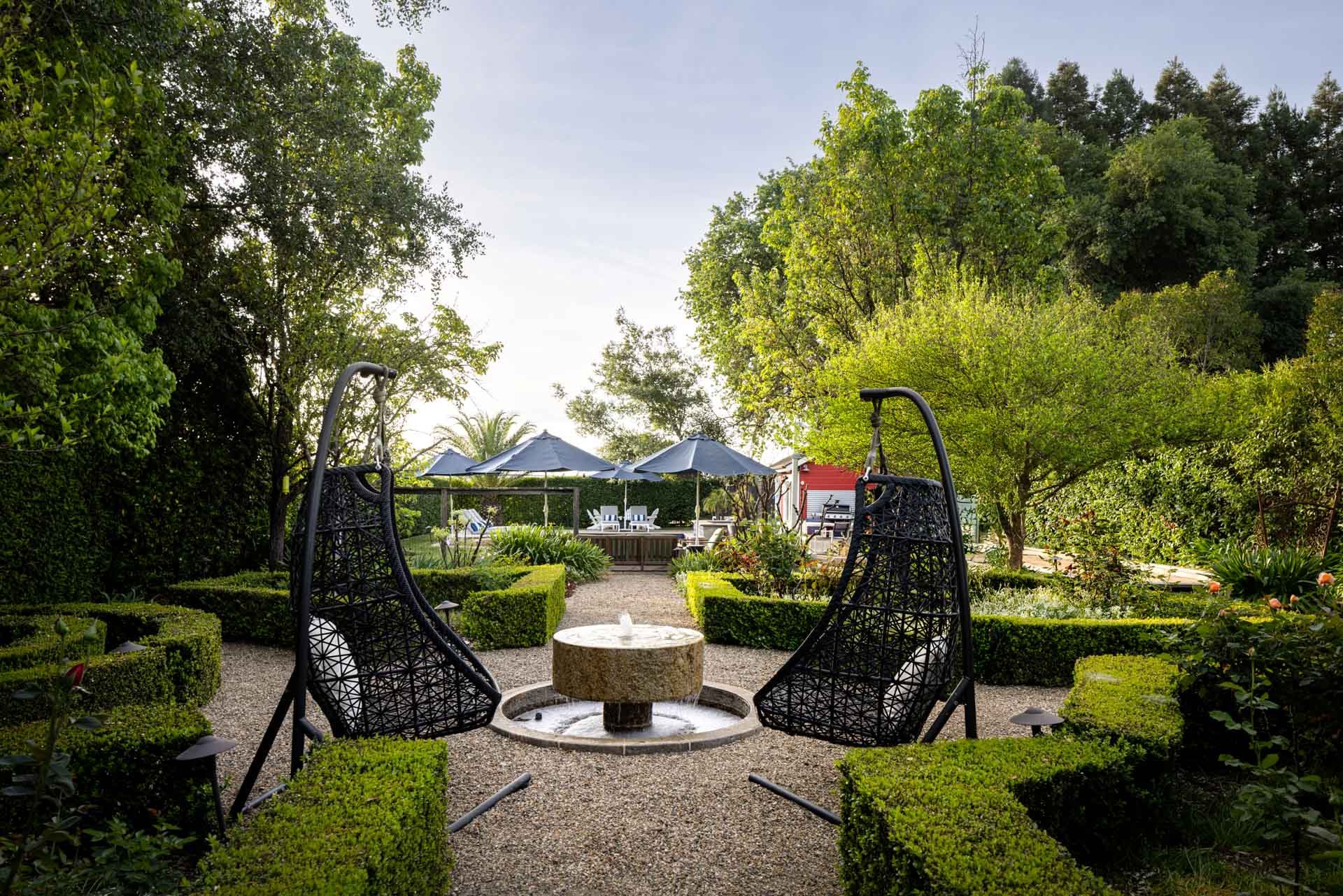 A backyard garden with green trees and shrubs, a gravel path, a small fountain in the center, two hanging chair swings, and a patio area with umbrellas and chairs in the background.