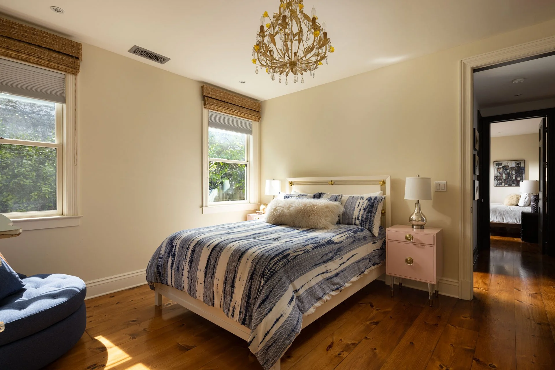 A bedroom with two windows, a bed with blue and white striped bedding, a pink nightstand with a lamp, and a chandelier. A doorway shows another room with a bed and artwork.