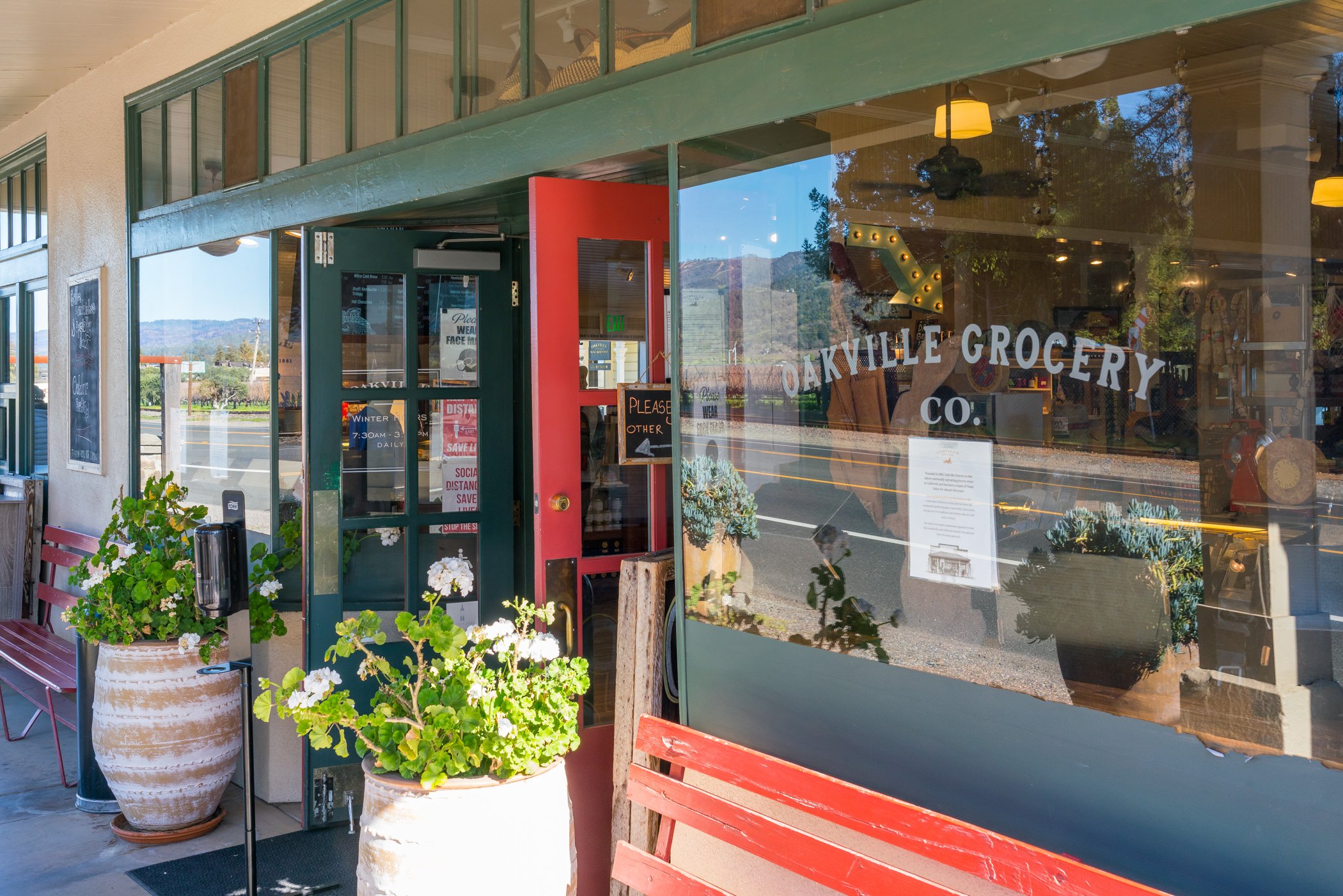 Exterior of Oakville Grocery Co. with large window, red door, potted plants with white flowers, benches, and sidewalk signs, reflecting trees and road in the window.