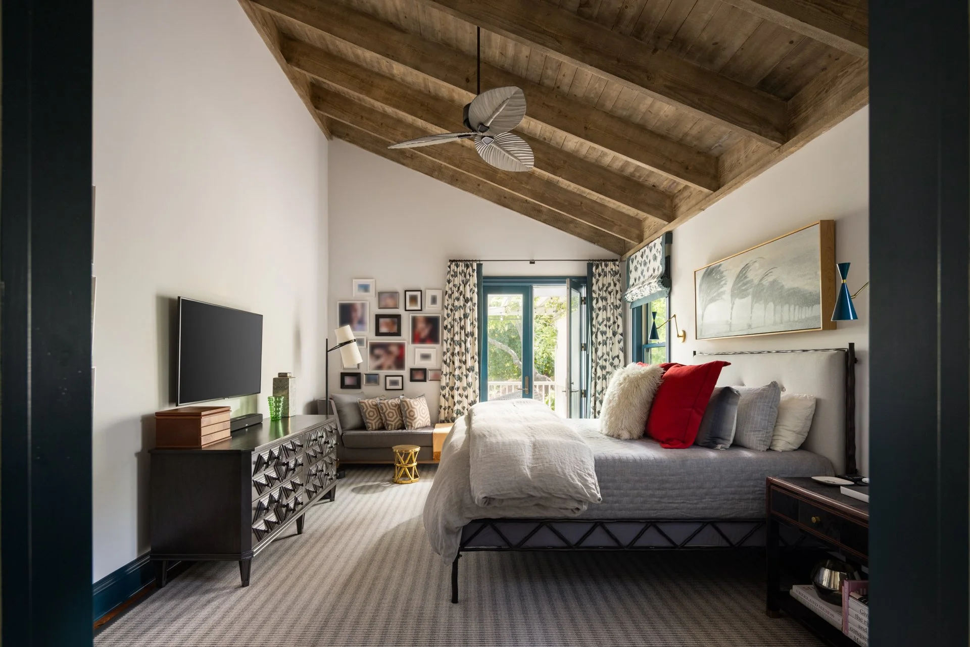 A bedroom with a sloped wooden ceiling, a bed with white and red pillows, a large artwork above the headboard, sliding glass doors leading to a balcony, a black dresser with a TV on top, and a sitting area near the window with a gray loveseat and pat