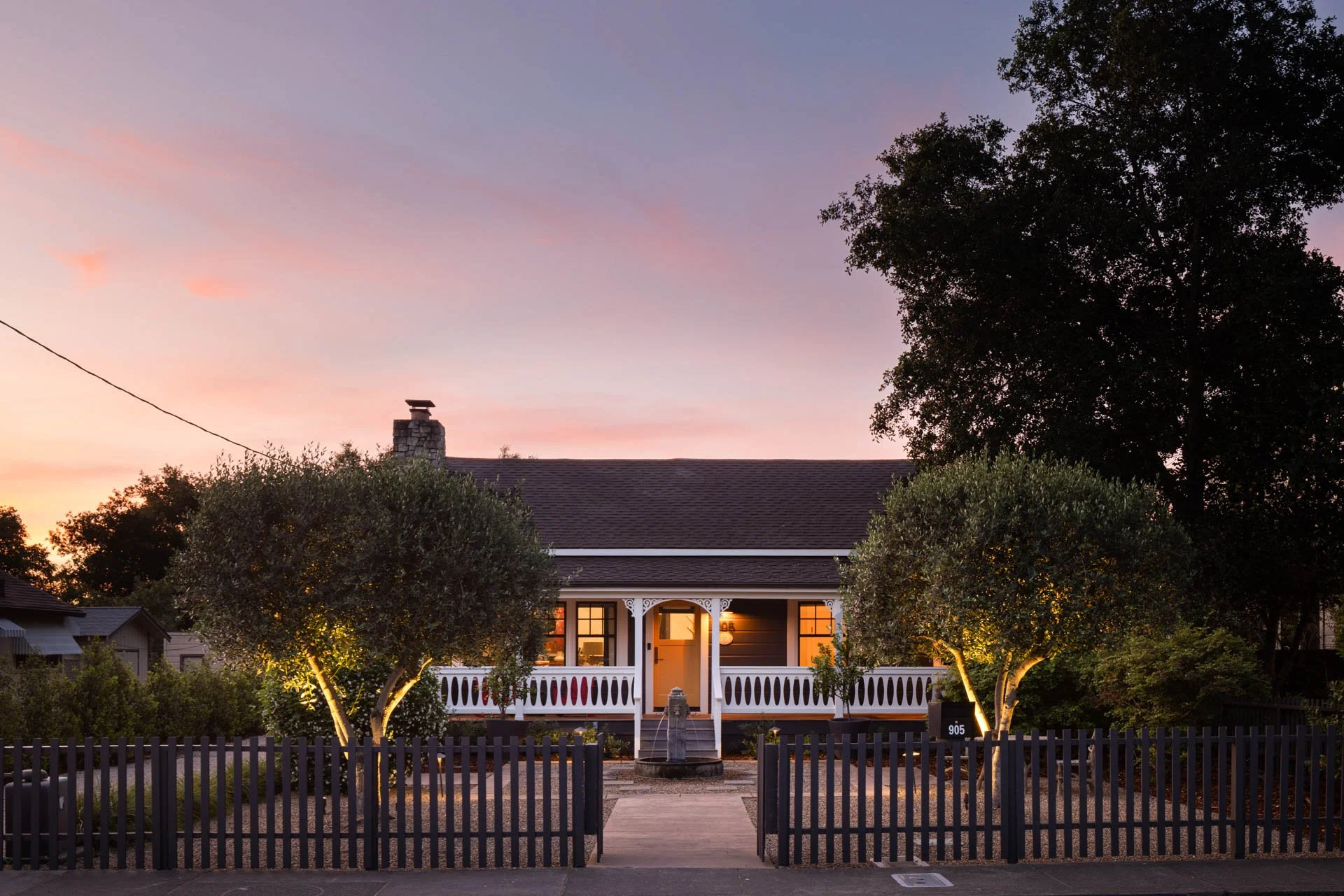 Front view of a house at dusk with warm lighting coming from the windows, two illuminated trees flanking the front porch, and a decorative fence along the sidewalk.