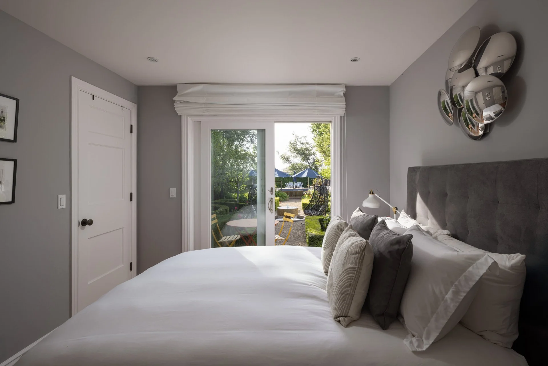Bedroom with gray walls, sliding glass door leading to a backyard patio with outdoor furniture and umbrellas, bed with gray headboard and white bedding, decorative pillows, and a wall mirror.