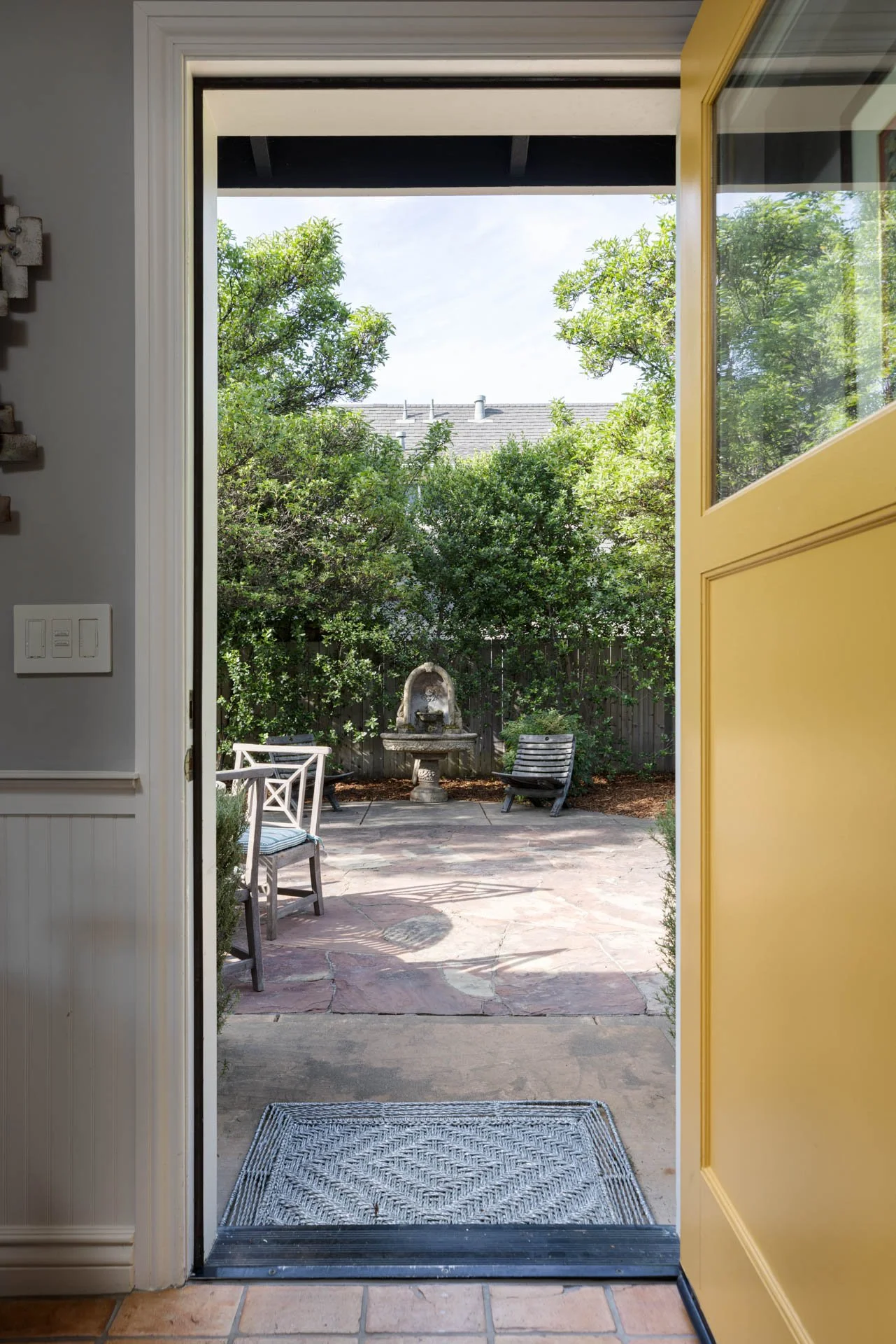 Open yellow door leading to a backyard patio with outdoor furniture, a fountain, and green trees.