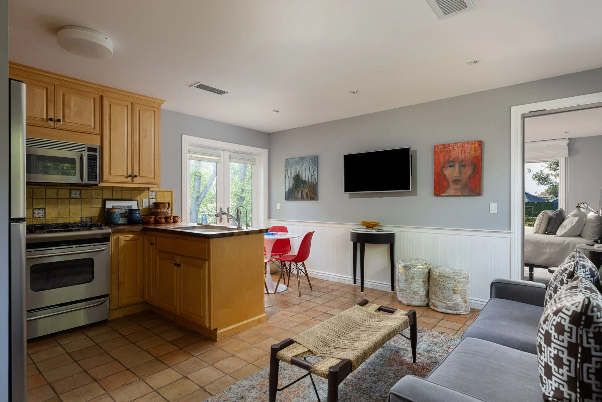 Open-concept kitchen and living room with wooden cabinetry, tiled floor, small dining table with red chairs, wall-mounted TV, and artwork on the walls.