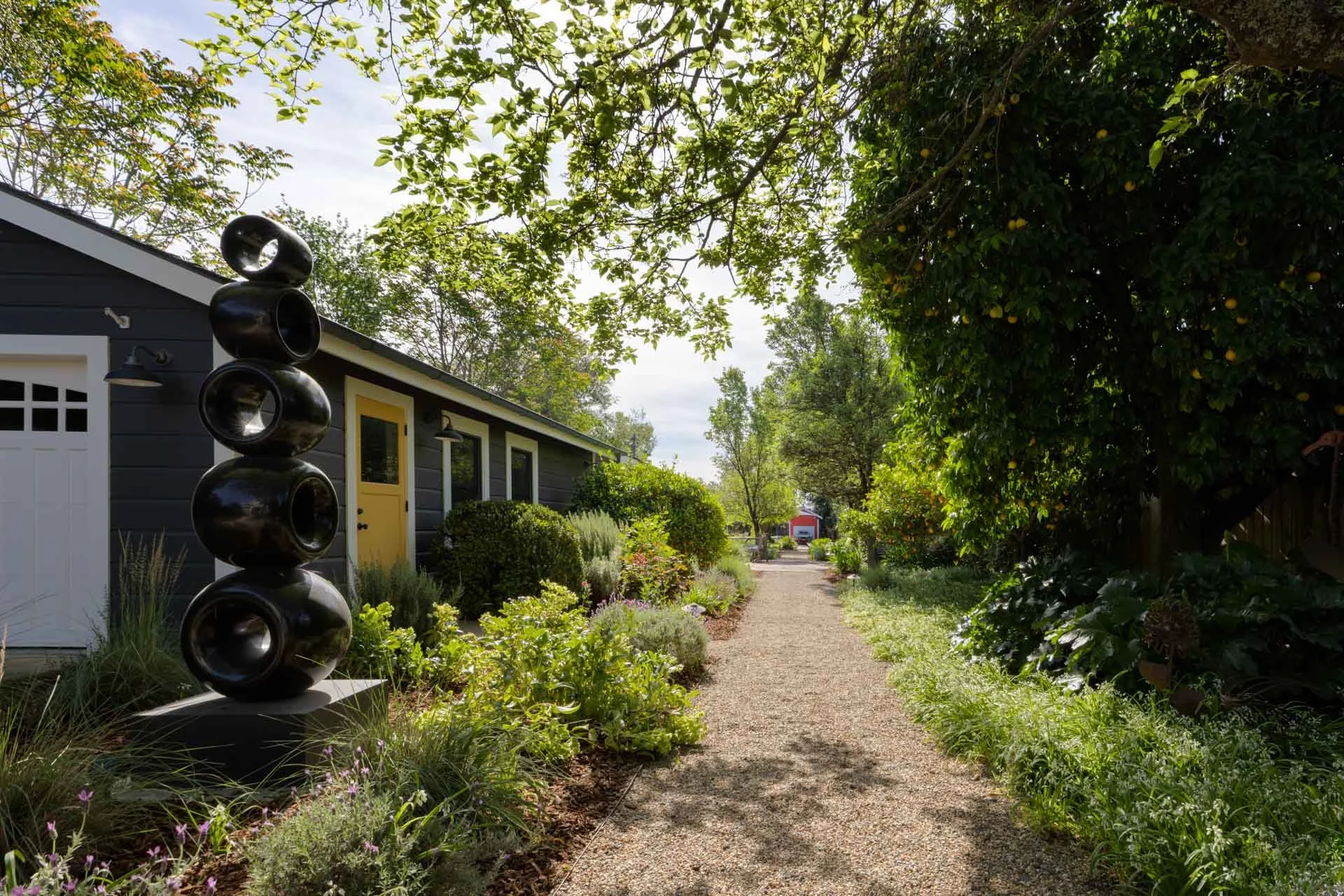 A gravel pathway runs through a lush garden, with a black sculpture of stacked rounded shapes on the left and a black house with a yellow door and window frames on the left side. Green trees and plants line the pathway, with a partly cloudy sky overh