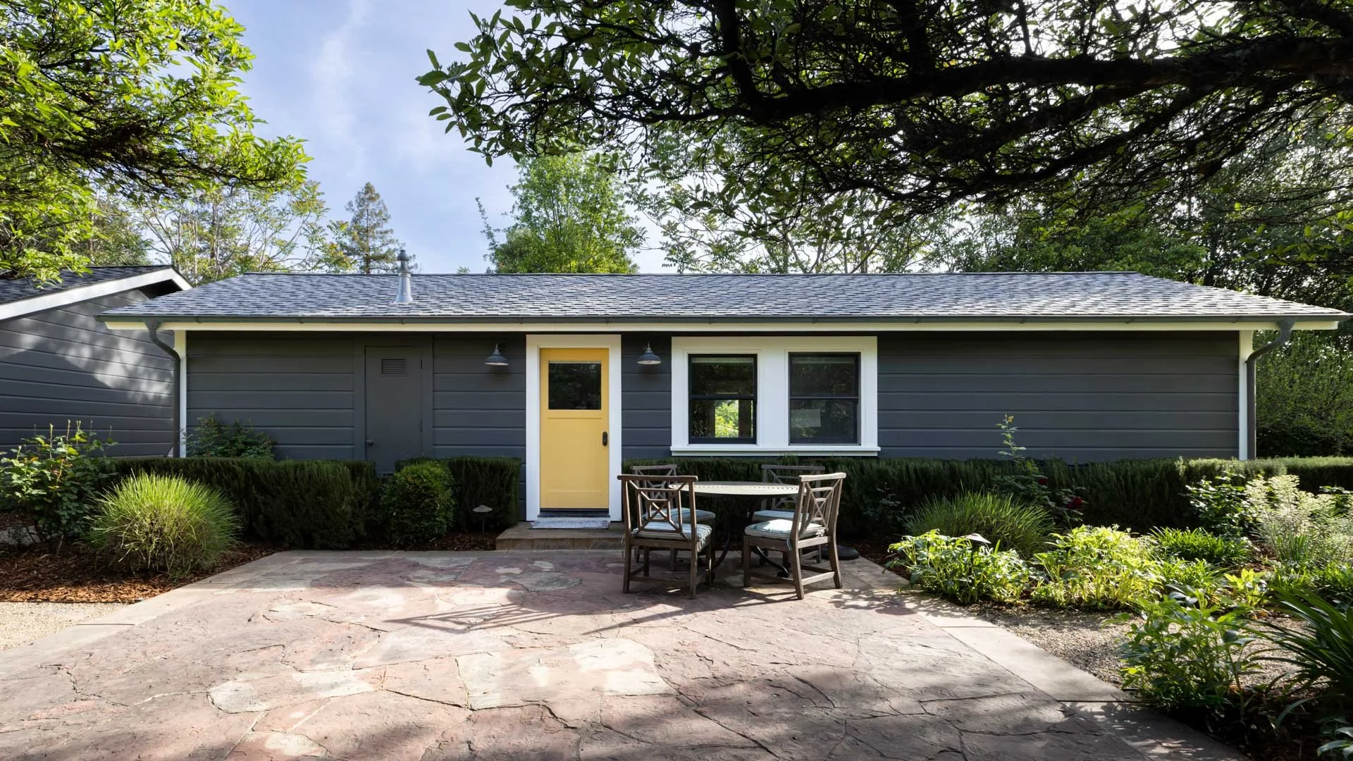 Front view of a gray house with a yellow door, two windows, and an outdoor patio with a round table and four chairs, surrounded by greenery and trees.