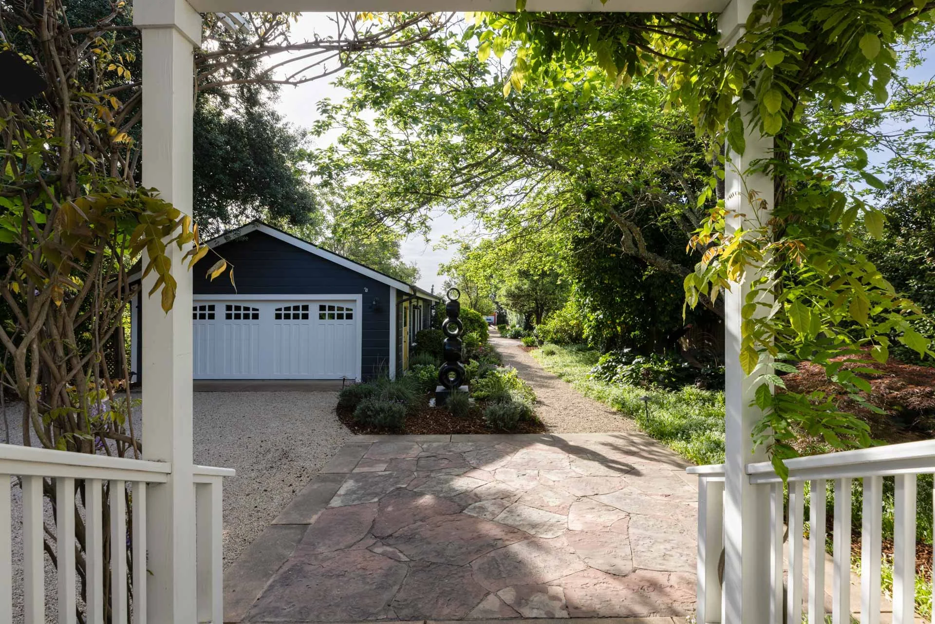 View through a garden gate showing a stone pathway, trees, and a blue garage with a white door.