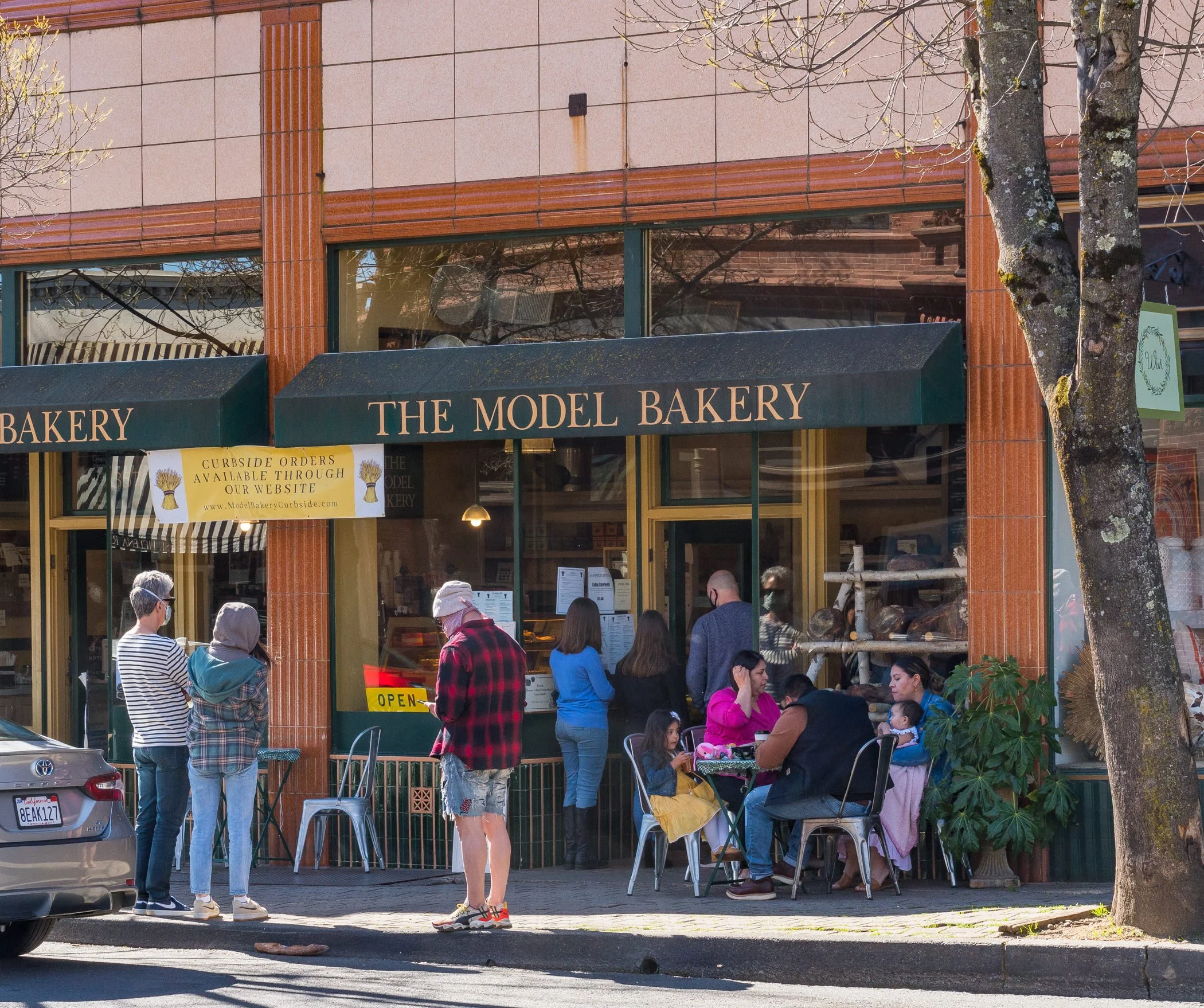 People standing outside and sitting at tables in front of a bakery called 'The Model Bakery' with a green awning and a sign indicating curbside orders are available.