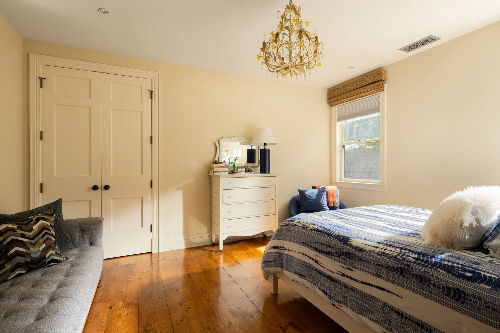 A bedroom with a bed covered in a blue and white striped blanket, a white dresser with a small mirror and a lamp, a navy blue armchair with pillows, hardwood floors, a window with a bamboo shade, a beige wall, a closed double door, and a chandelier h