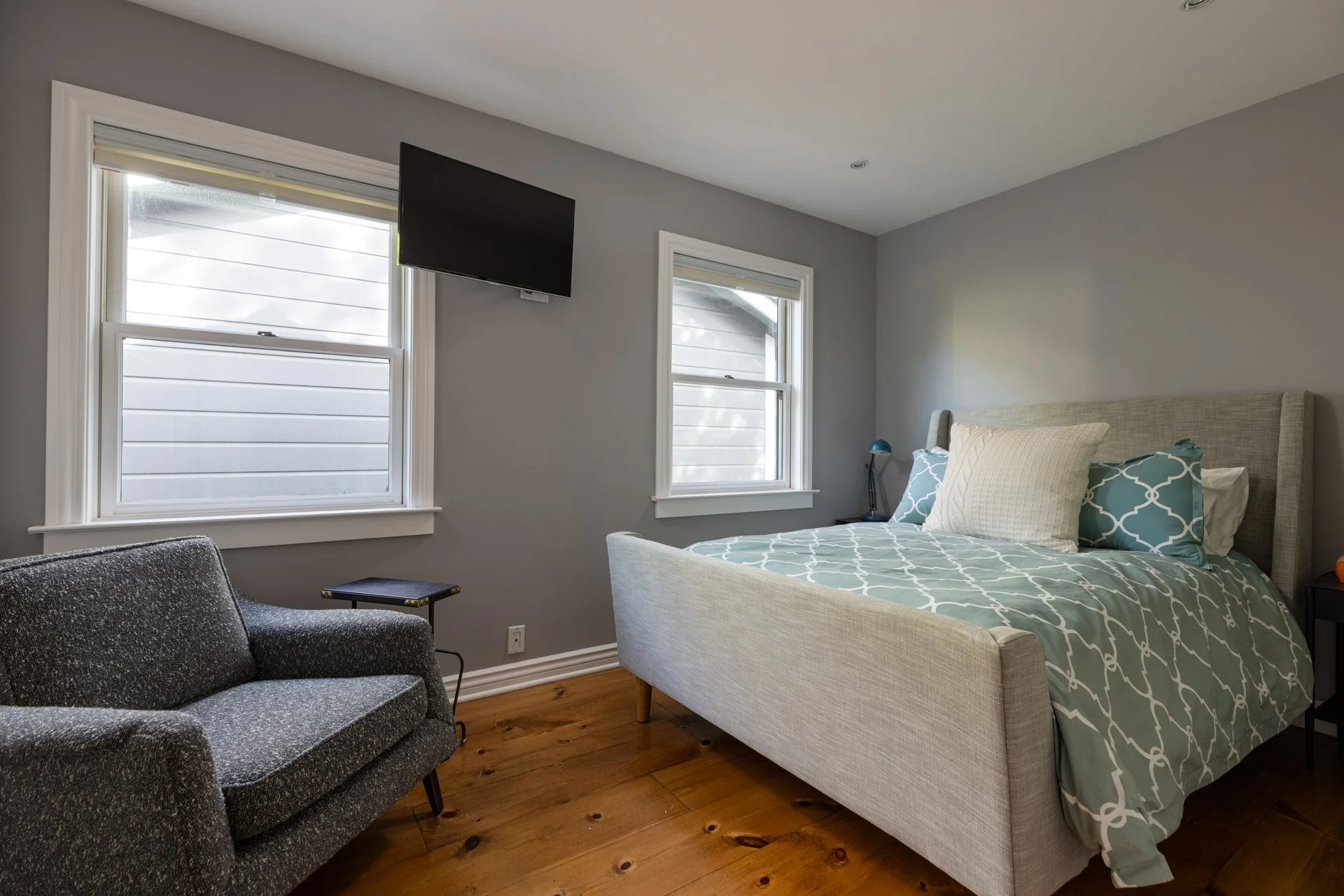 A bedroom with two windows, a gray armchair, a small side table, a wall-mounted flat-screen TV, a bed with a light-colored headboard, patterned bedding, and various pillows, with wooden flooring.