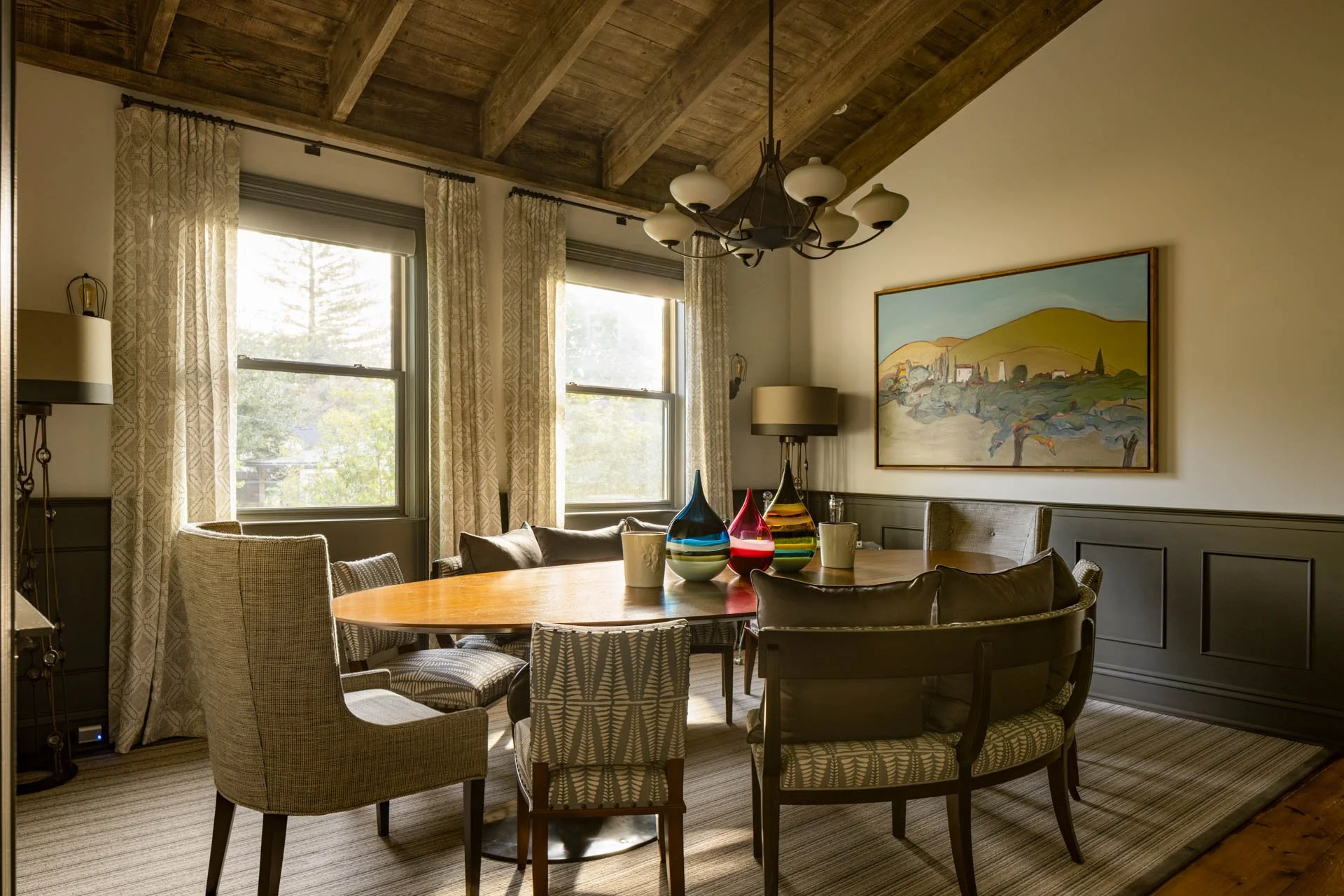 Dining room with wooden ceiling beams, framed landscape painting, multiple windows with patterned curtains, and a wooden table surrounded by mixed patterned chairs. Centerpiece includes colorful glass vases and mugs.