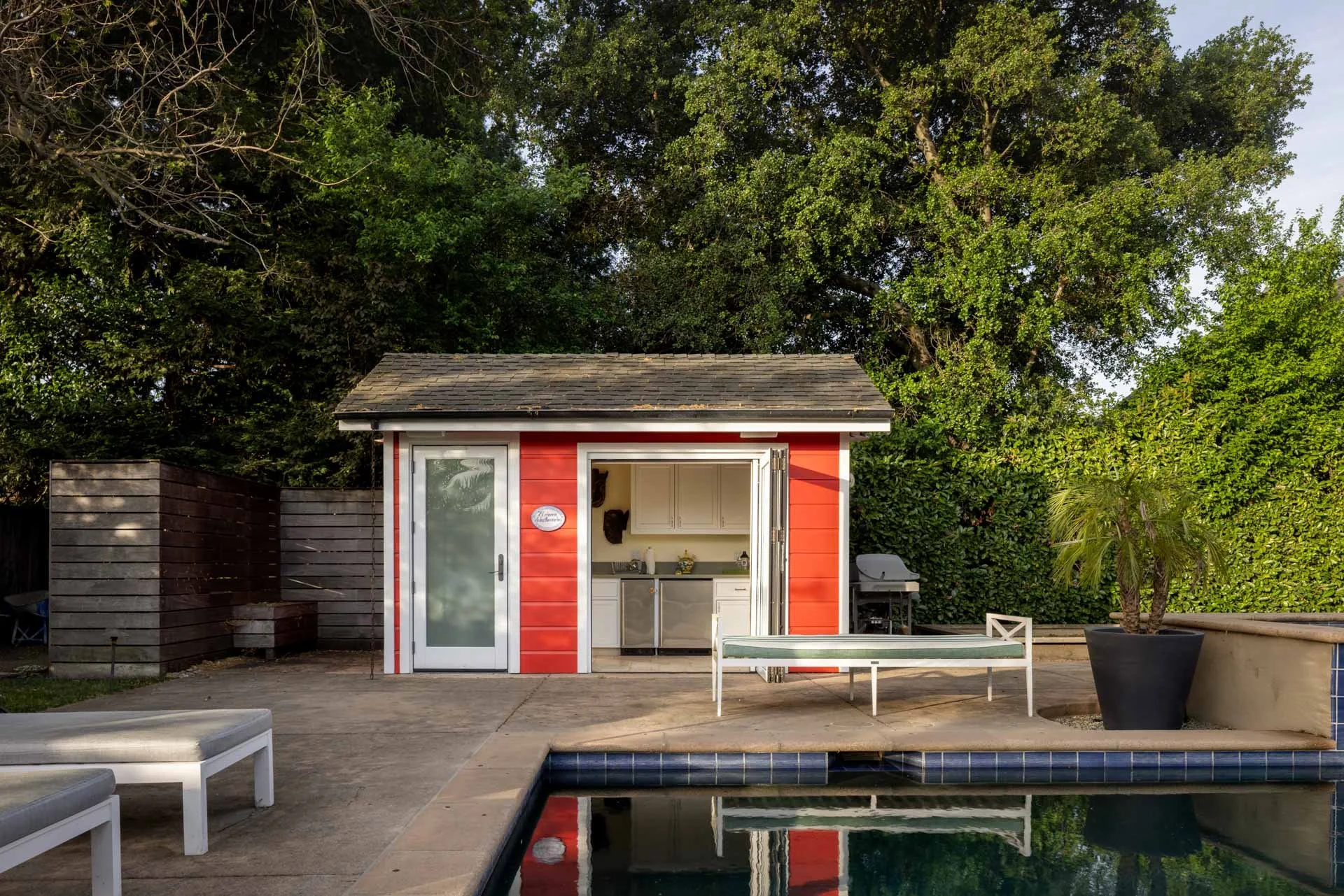 Backyard with a small red pool house, patio furniture, potted palm, outdoor grill, and swimming pool surrounded by green trees.