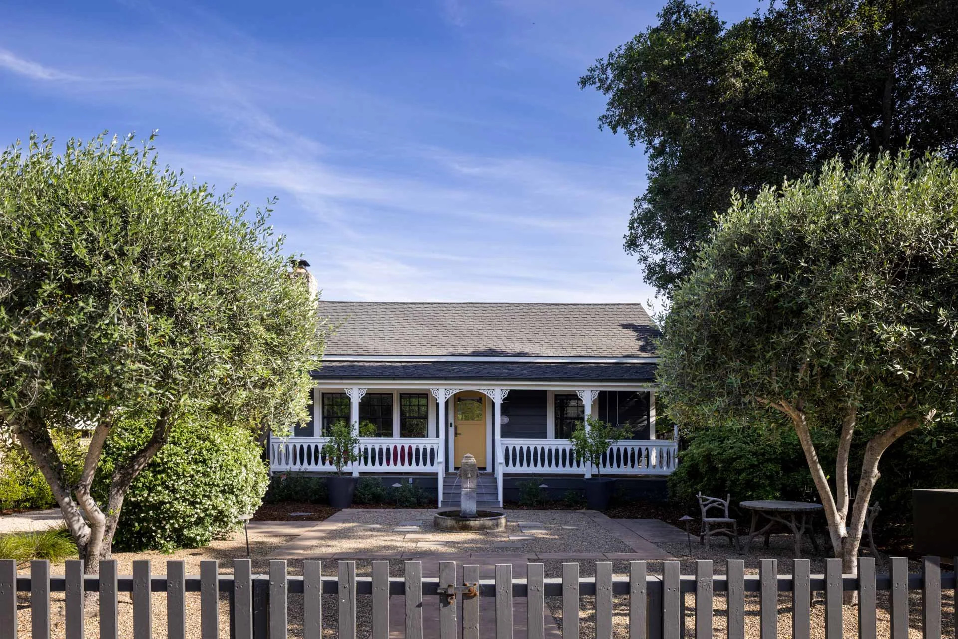 Front view of a small, dark gray house with white decorative trim, two trees on either side, a bench on the right, a fountain in front, and a gray picket fence in the foreground.