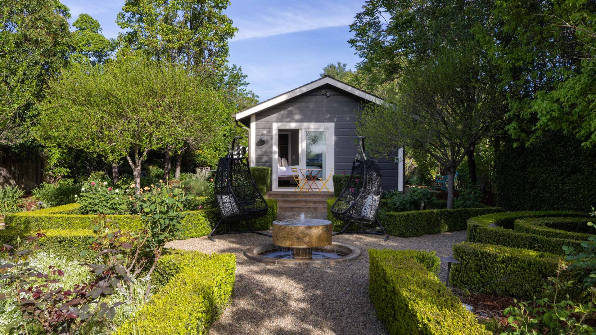 A backyard garden with a small gray house, a fountain in the foreground, and green trees and shrubs surrounding the area.