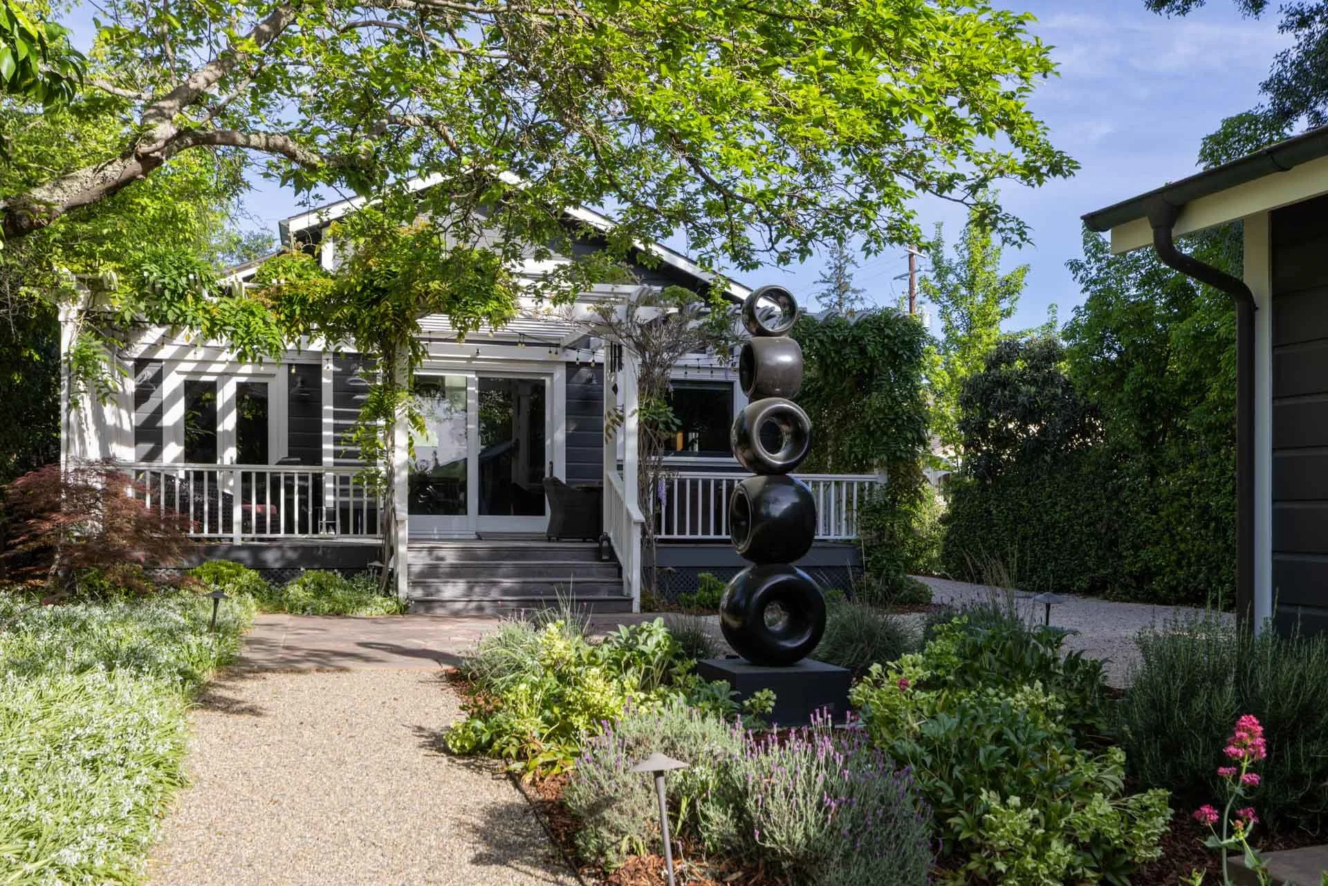 A garden with a pathway, various plants, trees, and a modern house in the background, with a black abstract sculpture
