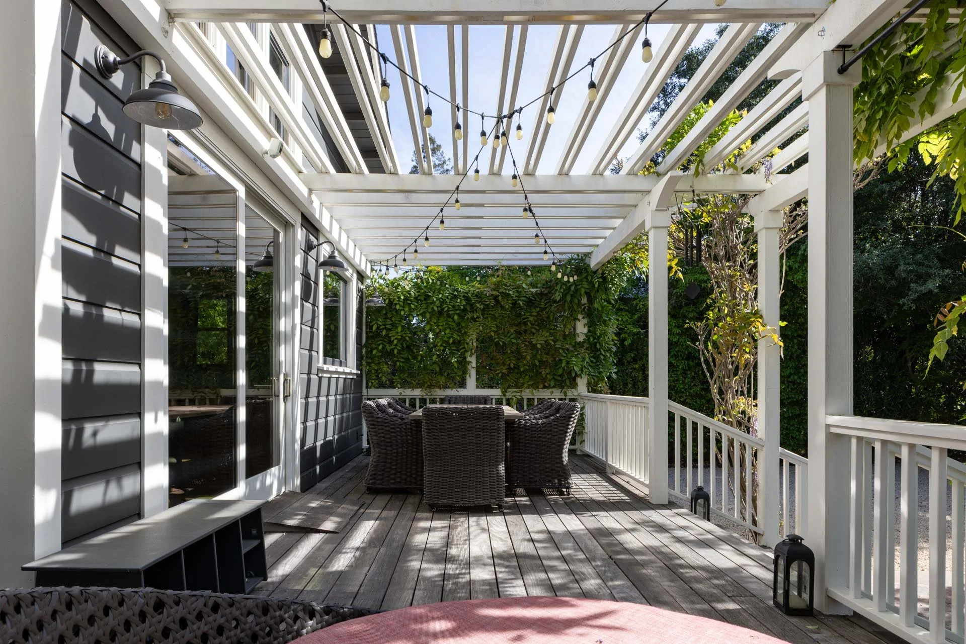 Outdoor patio with wooden deck, enclosed by white railing and privacy greenery, featuring a round table with six wicker chairs, string lights hanging from the white pergola, and decorative lanterns on the floor.