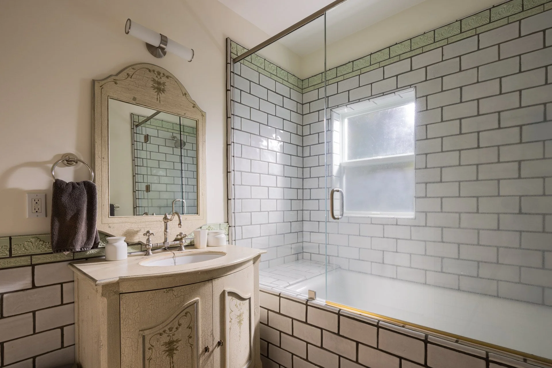 Bathroom with a vintage vanity, mirror, and a glass-enclosed shower with a window, beige walls, and decorative green tile border.