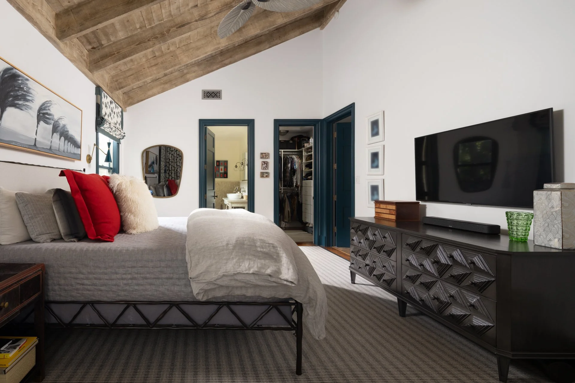 A bedroom with a bed, decorative pillows, a wall mirror, wall art, a dresser with items on top, and an open walk-in closet. There is a bathroom visible through an open door, and a flat-screen TV mounted on the wall opposite the bed. The room has wood