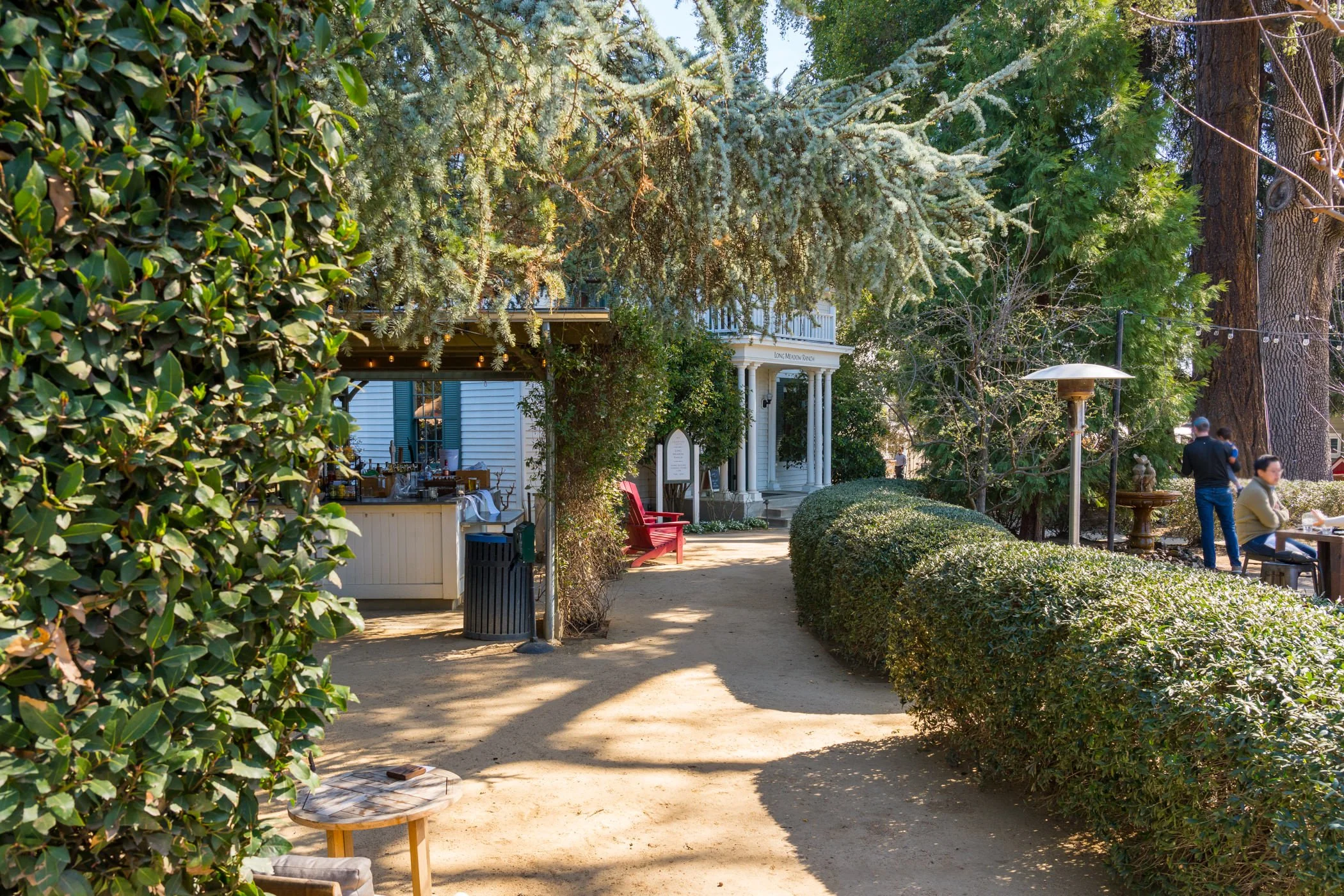 A peaceful outdoor garden scene with a sandy pathway leading to a white house with columns in the background. There are lush green trees and bushes on either side of the pathway, a red bench, and people sitting and standing near outdoor tables.
