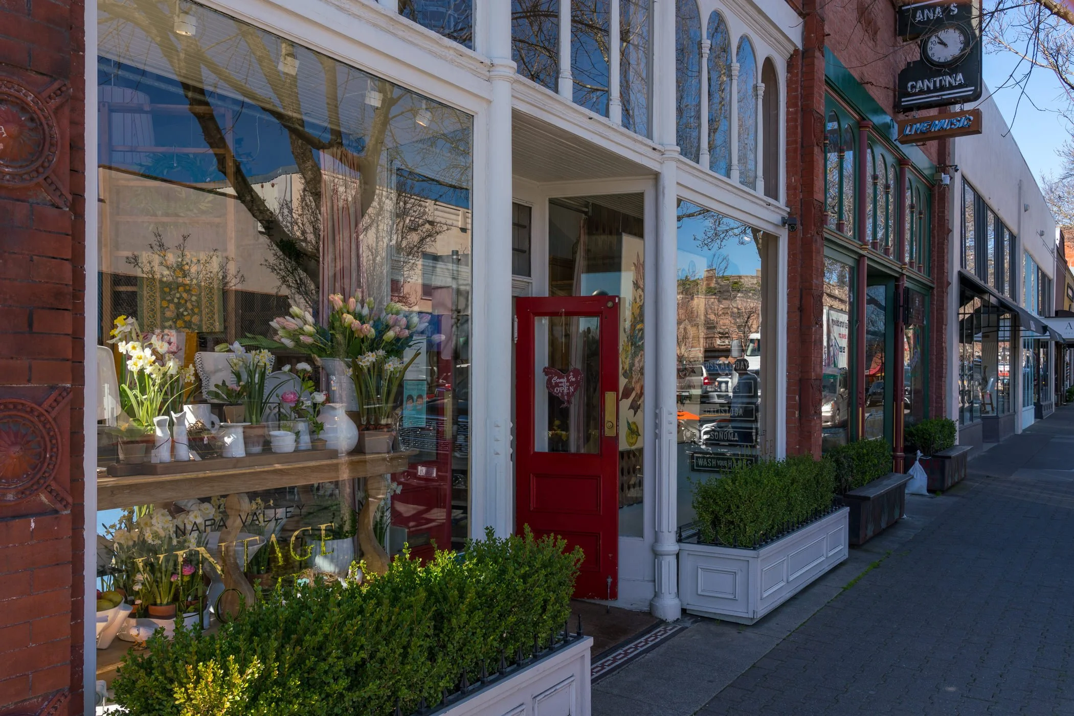 A storefront with large glass windows displaying flowers and plants, a red door with a heart-shaped sign, outside planters, and neighboring shops on a city street.