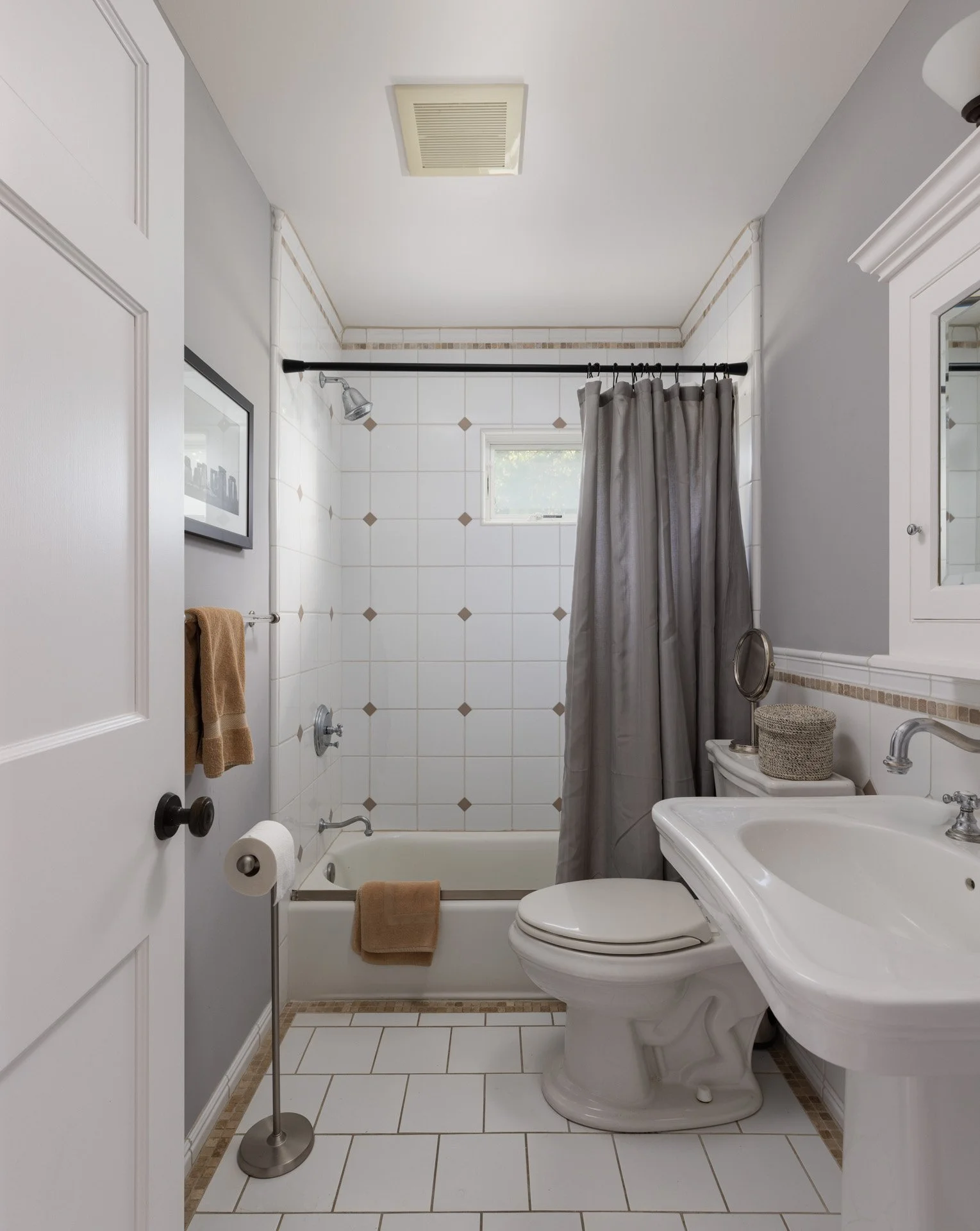 A small bathroom with a bathtub and shower, gray shower curtain, white tiles with beige accents, small window, white toilet, white sink, and a decorative mirror above the sink.