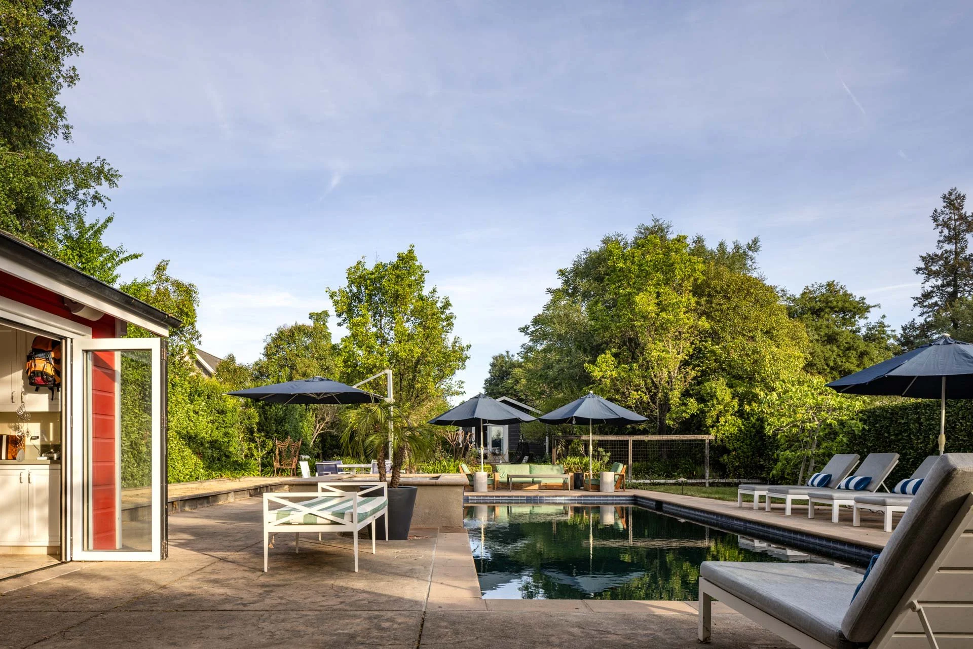 A backyard with a swimming pool, poolside lounge chairs with blue and white cushions, dark umbrellas, and lush green trees.