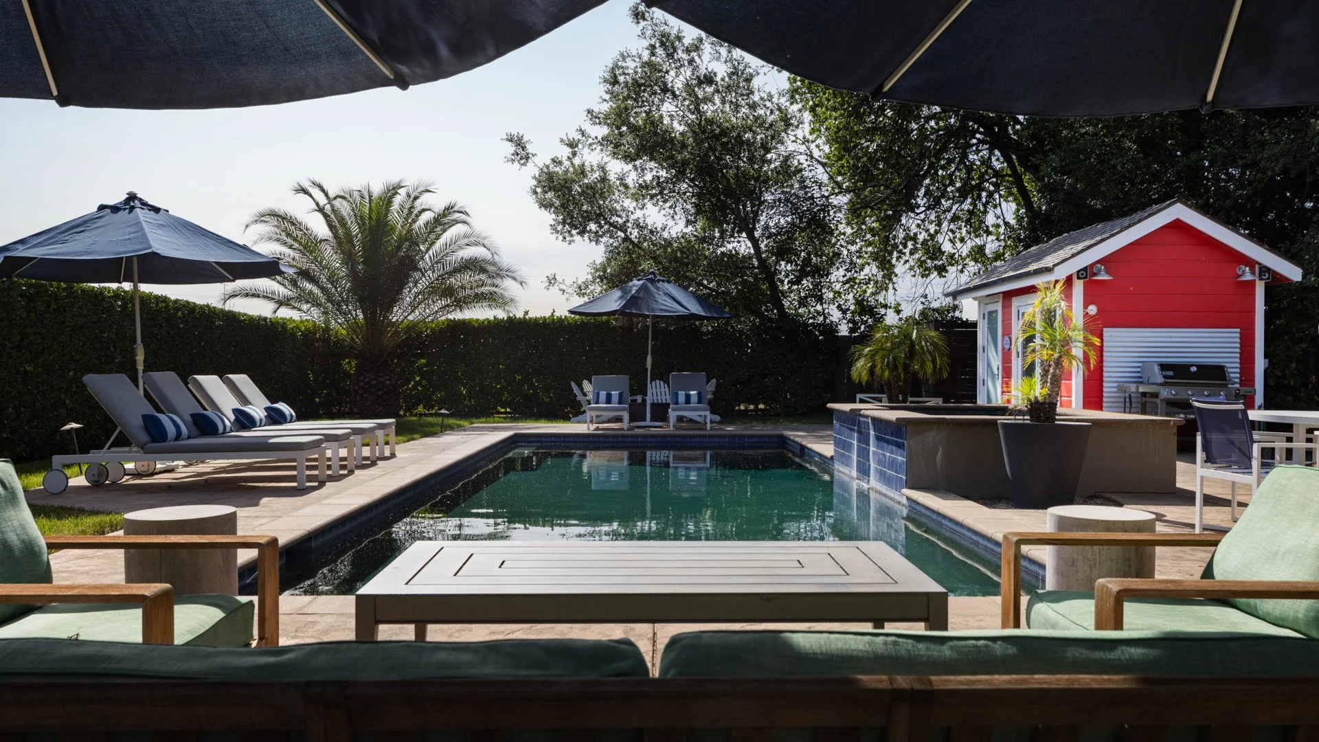 A backyard pool area with lounge chairs, umbrellas, a red pool house, potted plants, and outdoor furniture surrounded by trees and greenery.