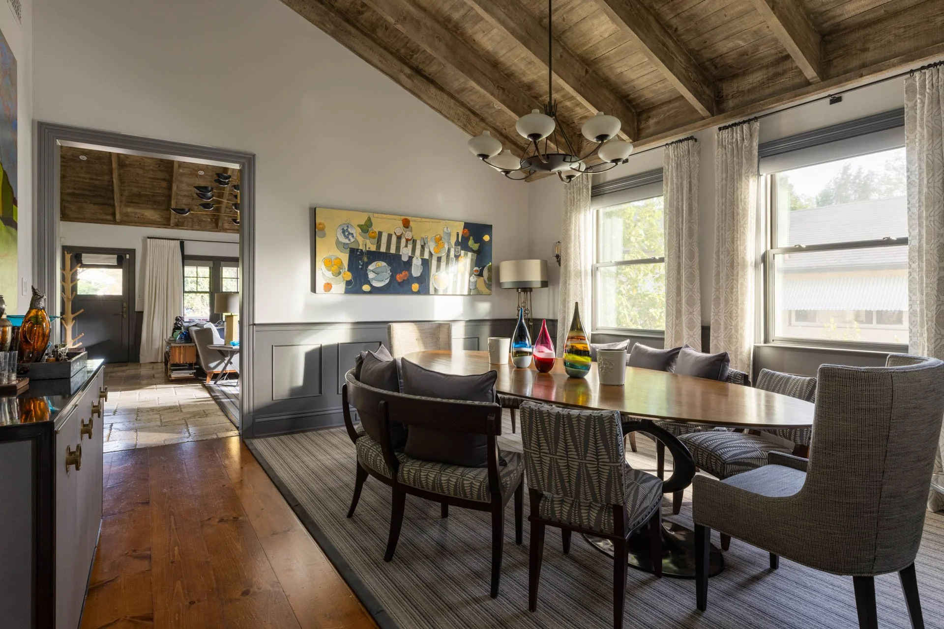 A dining room with a wooden oval table, surrounded by different patterned and solid-colored chairs, and decorative glass vases in the center. Large windows with patterned curtains let in natural light. A picture hangs on the wall, and a chandelier is