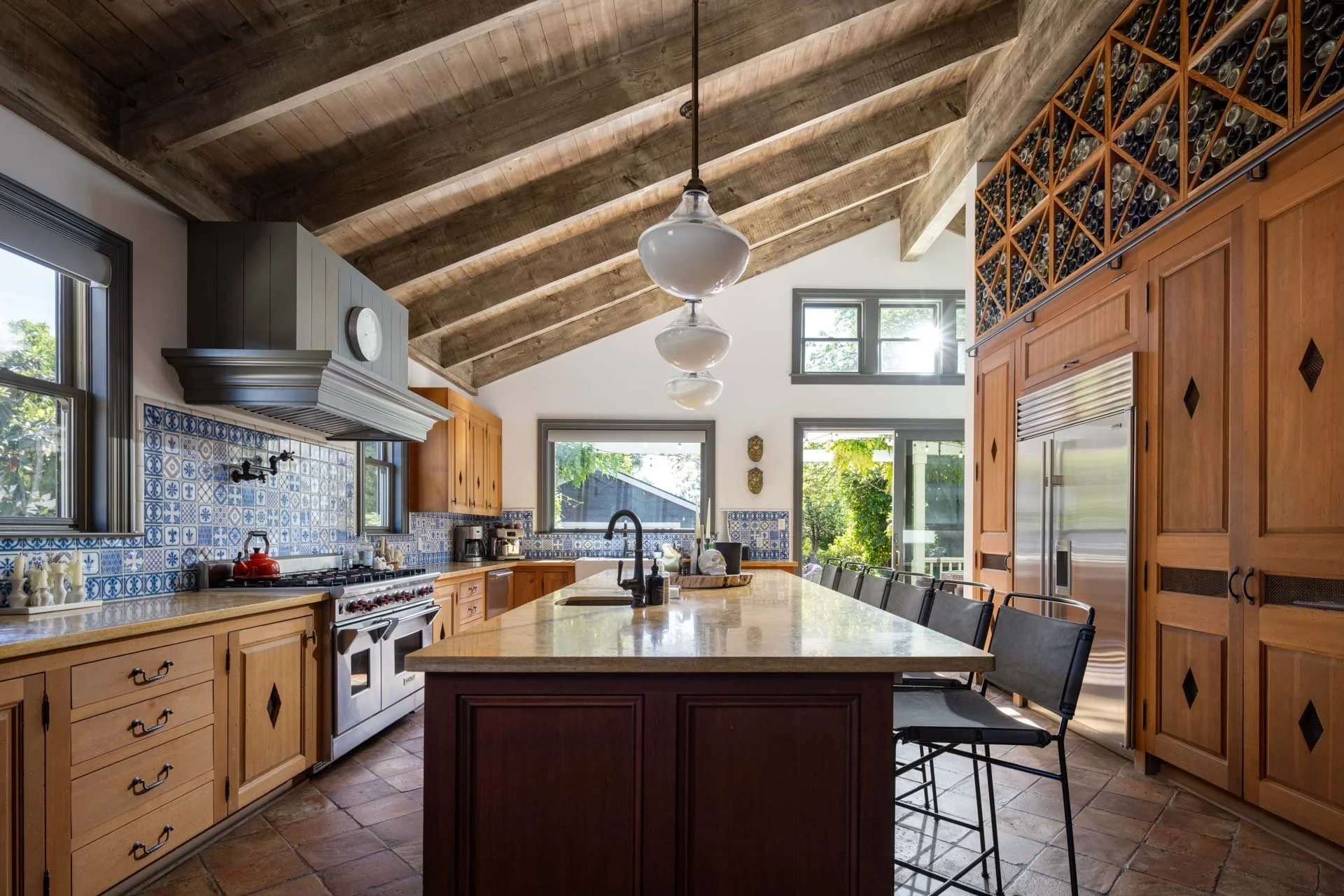 Kitchen with wooden cabinets, a central island, a stove with a metallic hood, blue patterned tile backsplash, and large windows letting in natural light.