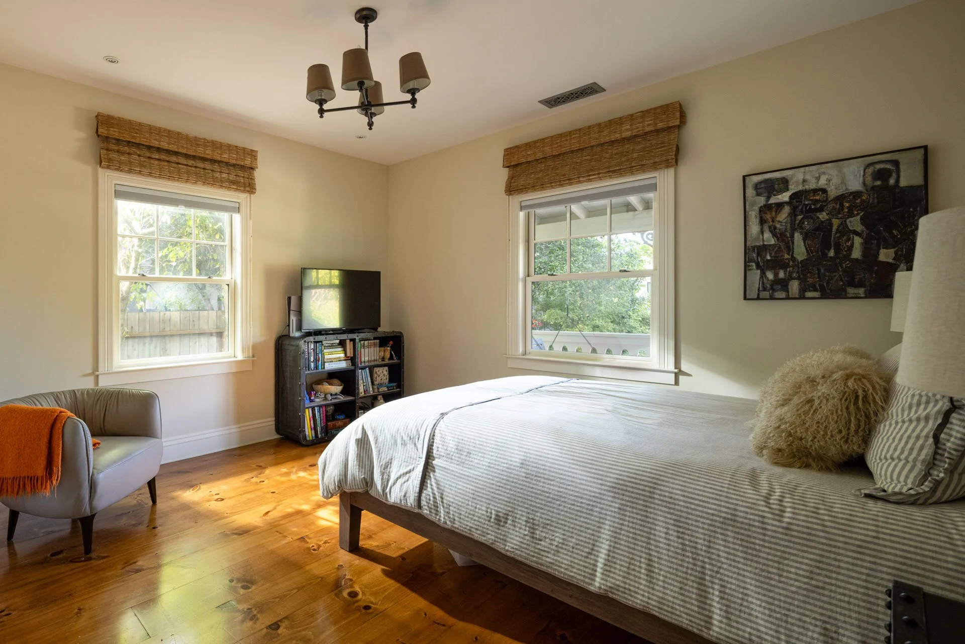 A bedroom with two windows, a bed with striped bedding and fluffy pillow, a chandelier, a small armchair with an orange blanket, and a TV on a black shelf with books.