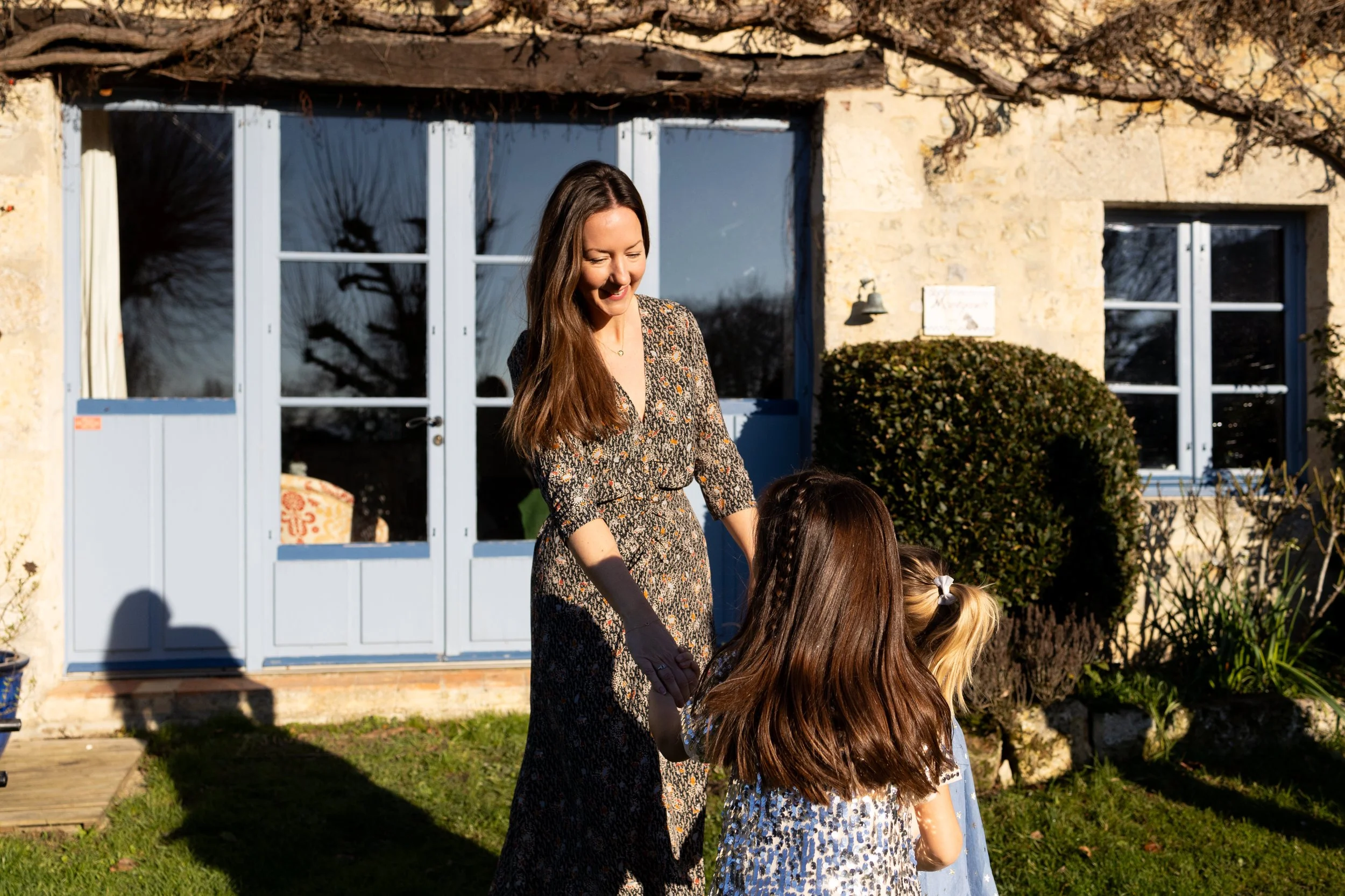 A woman with long brown hair wearing a patterned dress smiling and holding hands with two young girls with brown and blonde hair, standing outside in front of a stone house with white-framed windows and a door, sunny day with clear shadows.