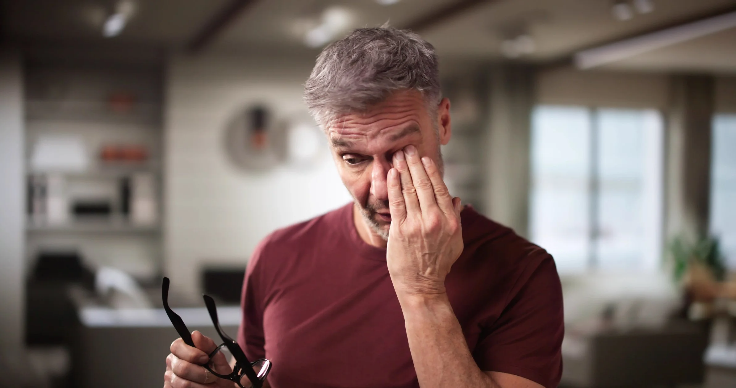 A middle-aged man with gray hair and a beard is rubbing his eye with his right hand while holding a pair of black glasses in his left hand due to blurred vision