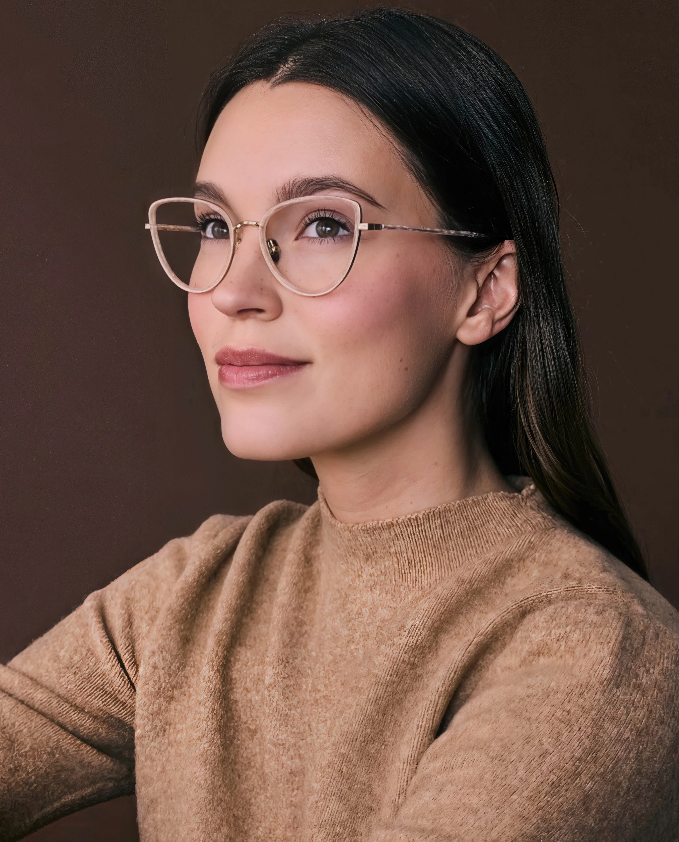 Close-up portrait of a young woman with dark hair, wearing glasses and a beige sweater, looking to the side against a brown background.
