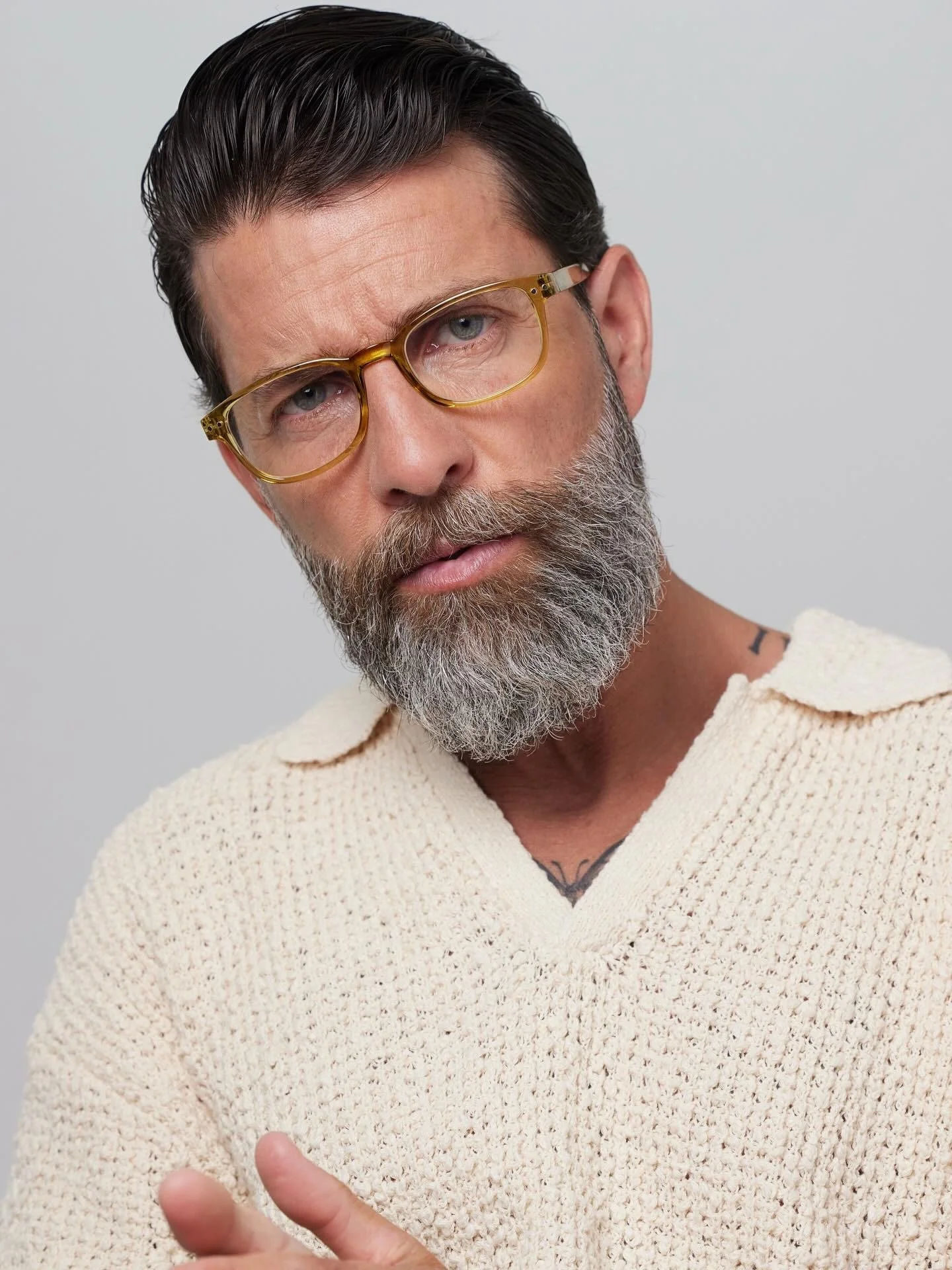 A middle-aged man with dark hair, a gray beard, and yellow glasses looks directly at the camera. He is wearing a cream-colored knit sweater and has a tattoo visible on his chest, partially covered by his sweater.