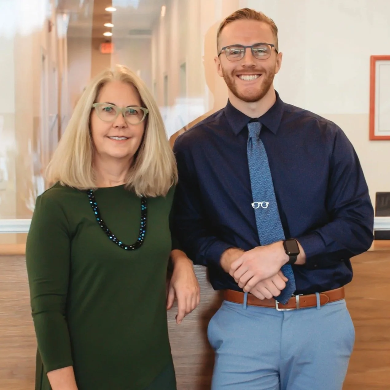 A woman wearing a green top and glasses standing next to a man in a dark shirt, light-colored pants, and glasses, both smiling indoors.