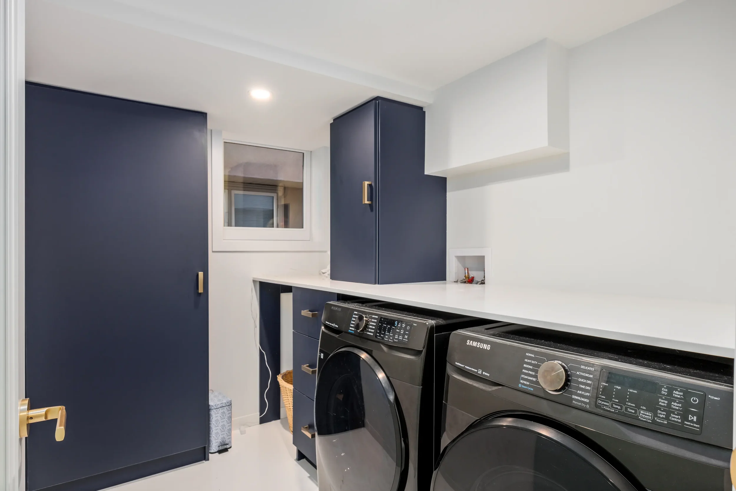 A laundry room with dark blue cabinets, a white countertop, a window, a Samsung washer and dryer, and a small basket on the floor.