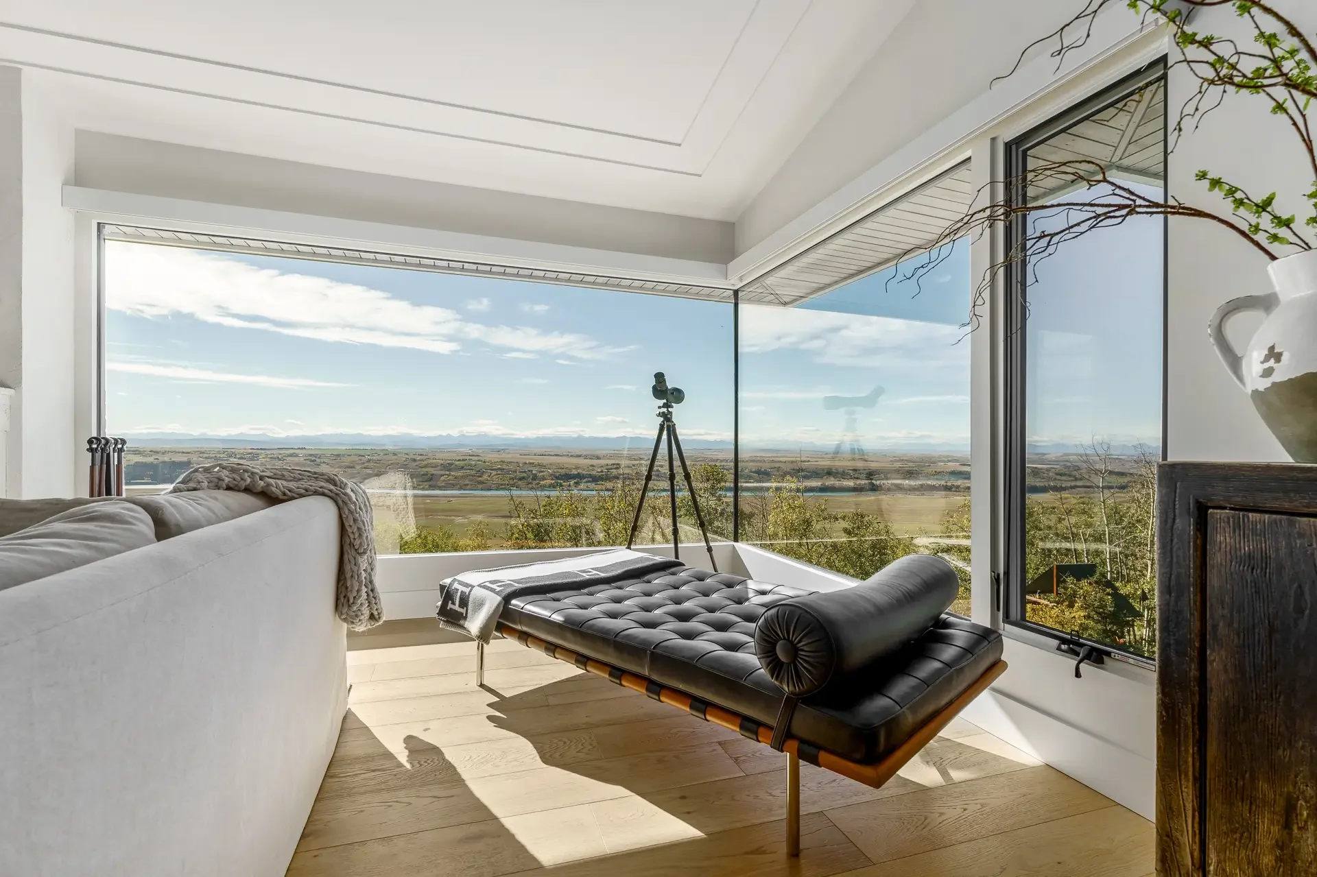 Interior view of a modern living room with large floor-to-ceiling windows showing a scenic landscape of fields and mountains, with a telescope set up near the window and a black leather lounge chair. A white couch, decorative pillows, and a knitted t