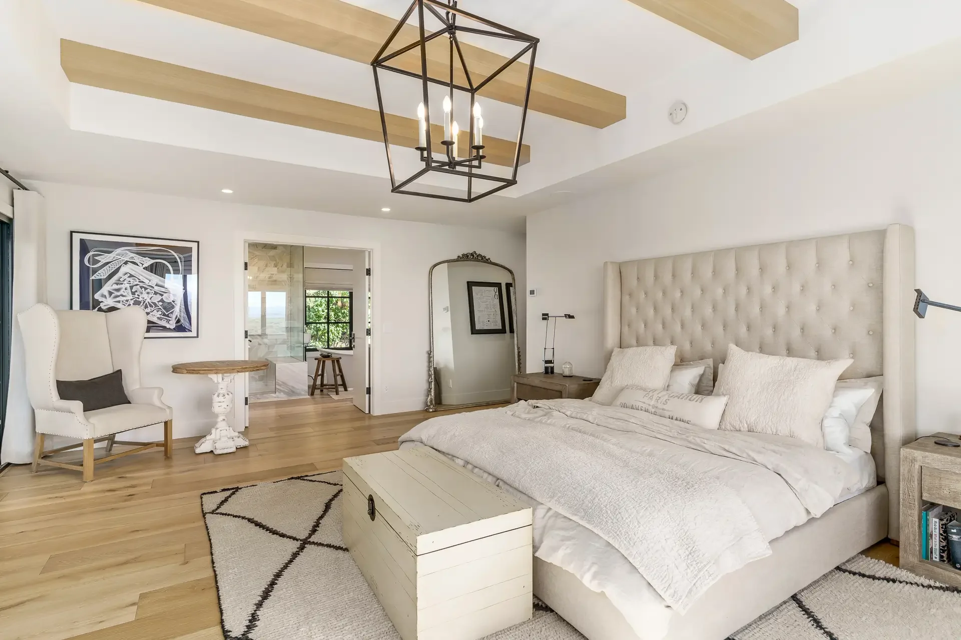 A bedroom with a large, tufted, beige headboard, white bedding, and pillows. A white wooden chest at the foot of the bed. Light wood floors and a textured rug with black lines. A black metal chandelier with candle-style lights hangs from the ceiling.