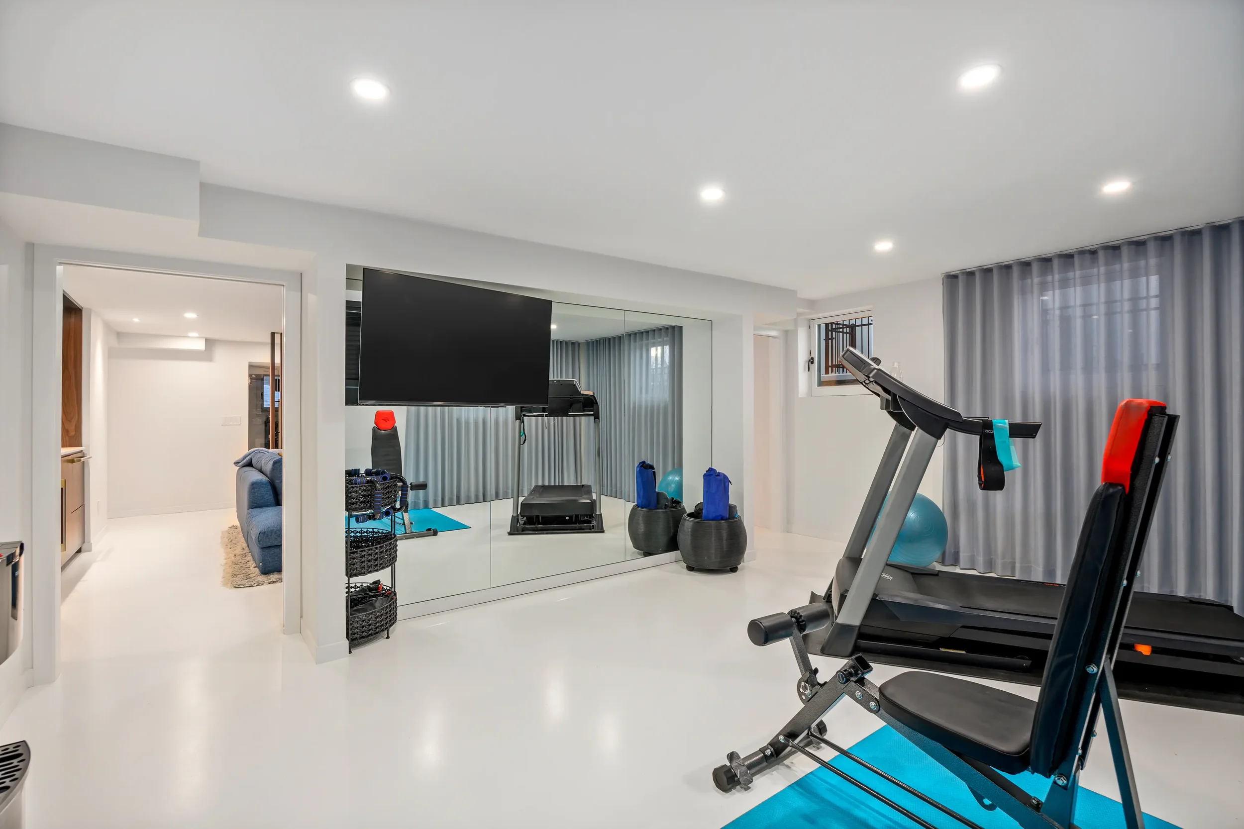 Home gym with treadmill, exercise equipment, large TV, mirrored wall, and gray curtains.
